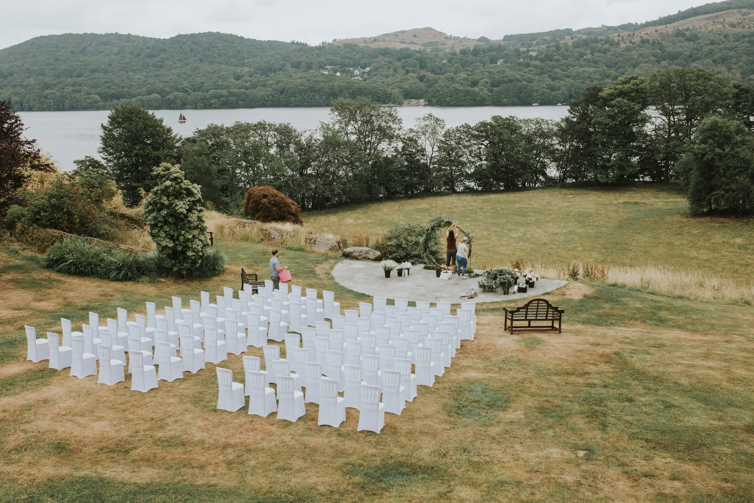 Preparation area for an outdoor wedding ceremony by a lake, with white chairs arranged facing a wooden arch, and scenic green hills in the background.