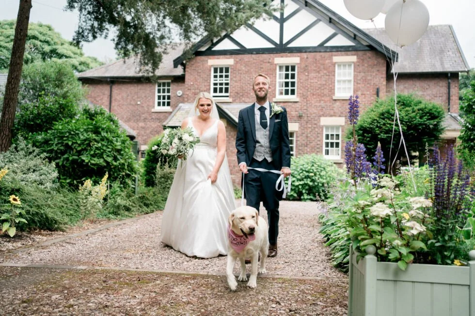 A bride and groom walking their dog outside a brick house with lush greenery and flowers.