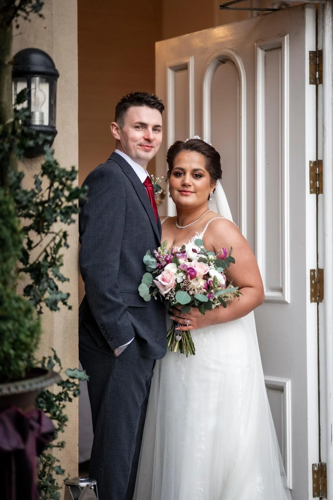 A bride and groom standing at the doorway of a house, posing for a photo during their wedding day. The groom is wearing a gray suit with a red tie, and the bride is in a white wedding dress holding a bouquet of pink, purple, and white flowers.