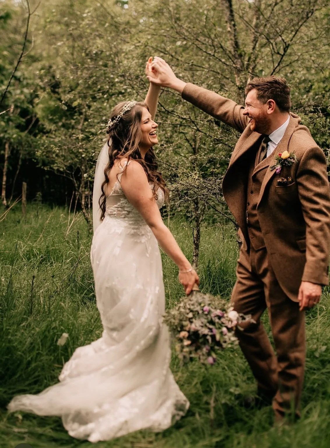 A bride and groom dancing outdoors in a lush, green area with trees, smiling and holding hands, with the bride holding a bouquet of flowers.
