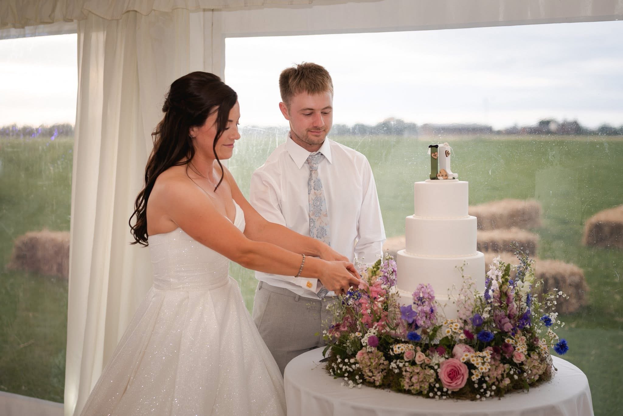 A bride and groom cutting a wedding cake together at their outdoor wedding celebration, with a table filled with colorful flowers in front of them and a scenic outdoor view through a window behind them.