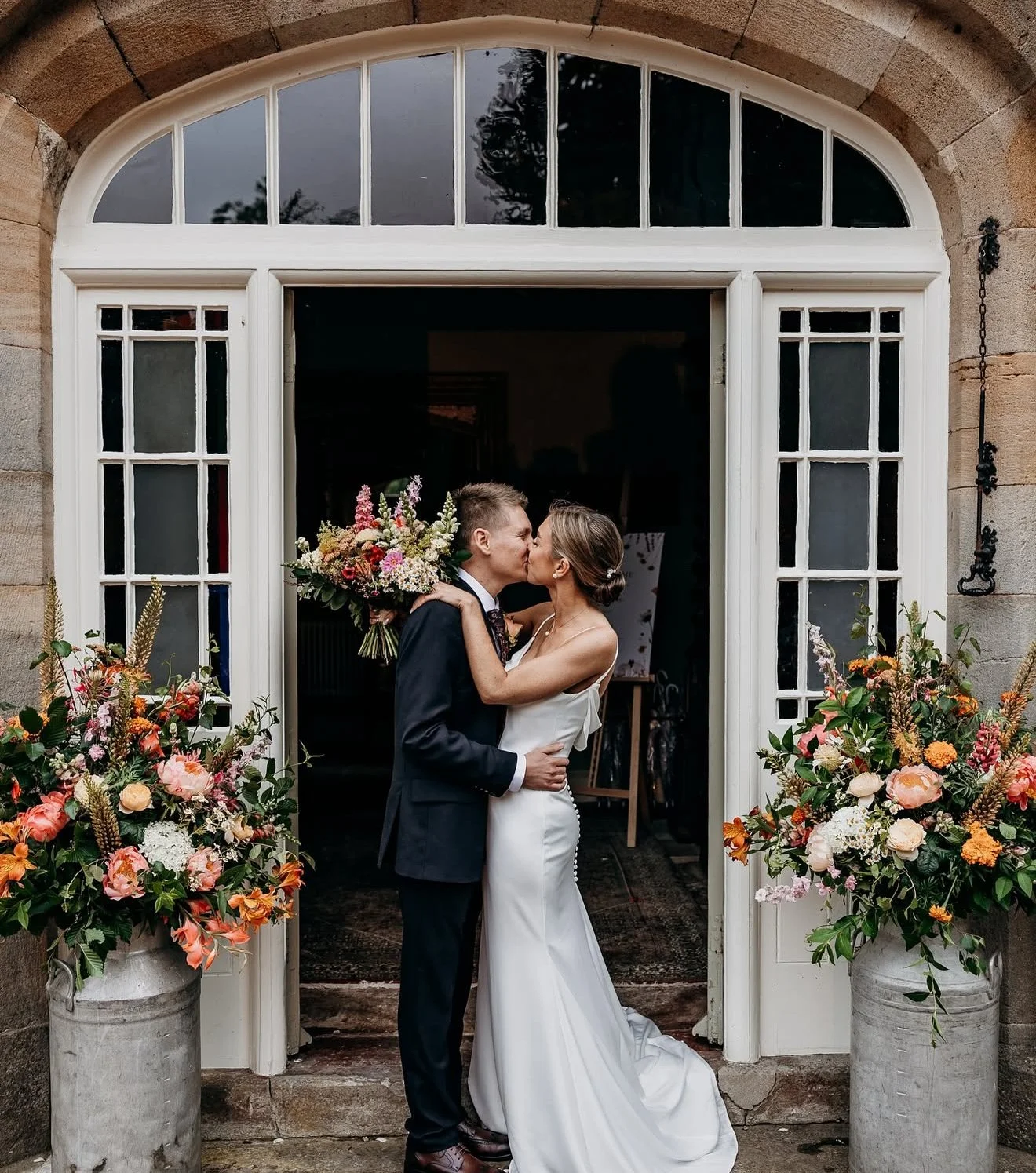 A bride and groom kiss in front of a building with floral arrangements on either side of an open door.