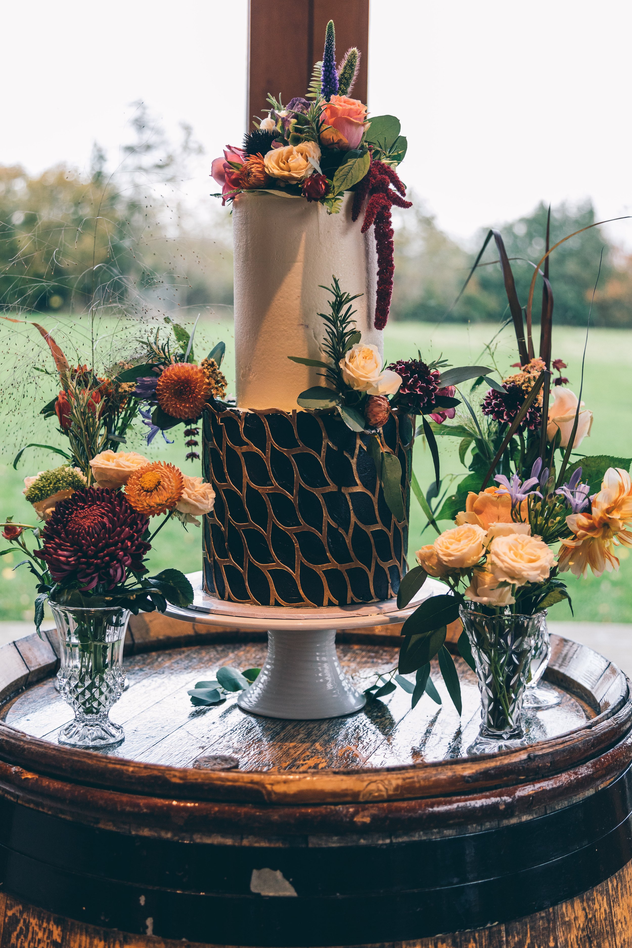 A tiered white wedding cake with gold pattern, decorated with flowers, surrounded by flower arrangements in crystal vases on a wooden table.
