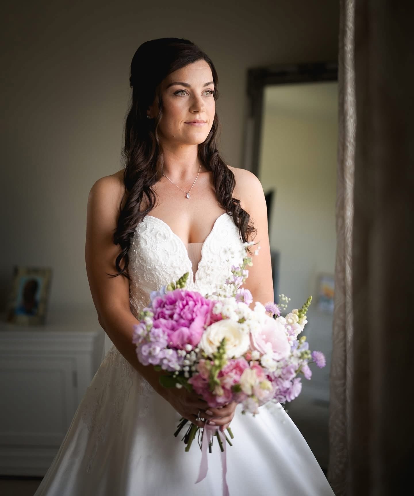 A young woman in a wedding dress holding a bouquet of pink, white, and purple flowers, standing indoors near a mirror.