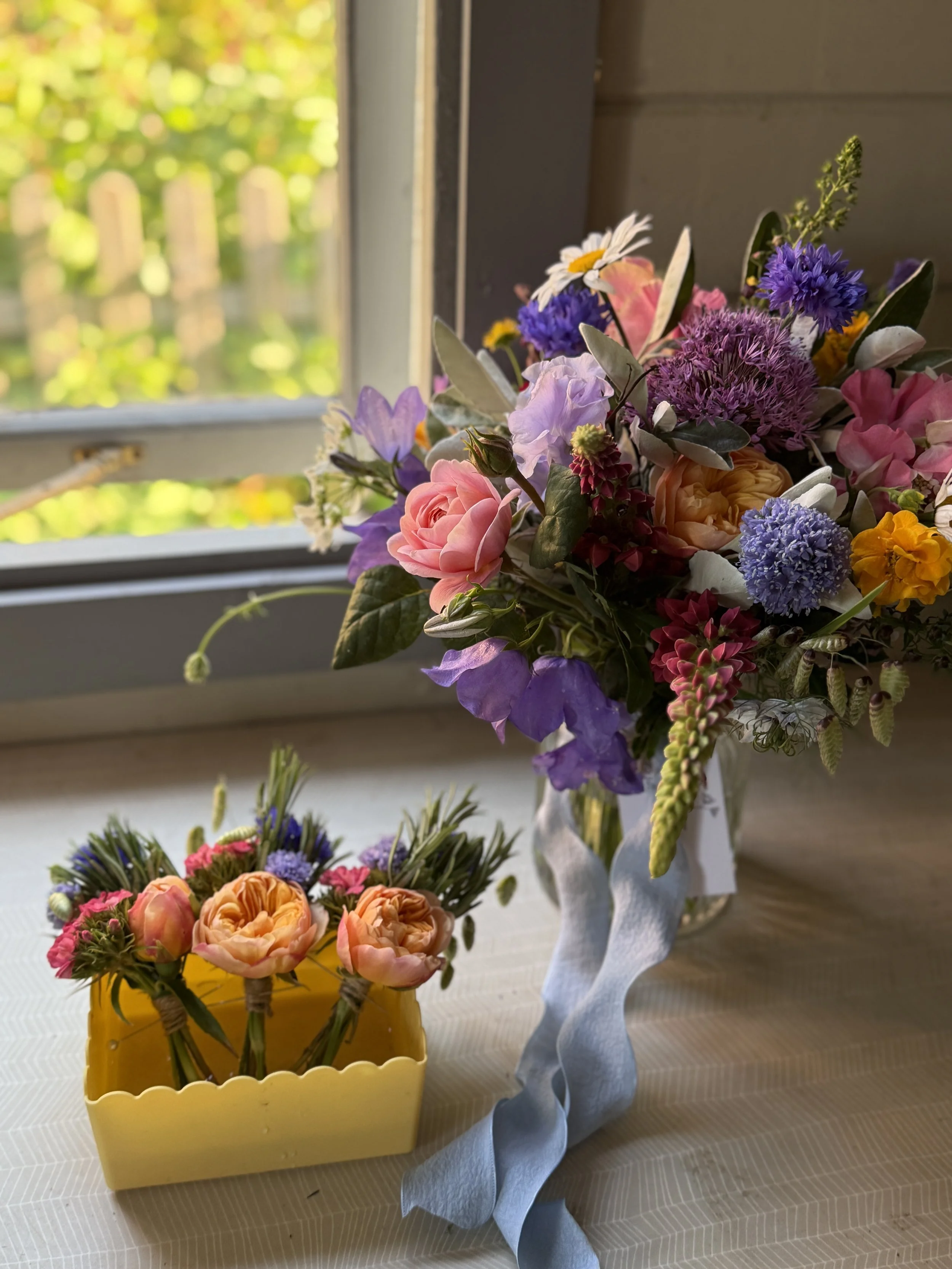 A colorful bouquet of flowers in a white cloth vase on a wooden table, with a window showing green foliage outside and a smaller yellow flower basket in the foreground.