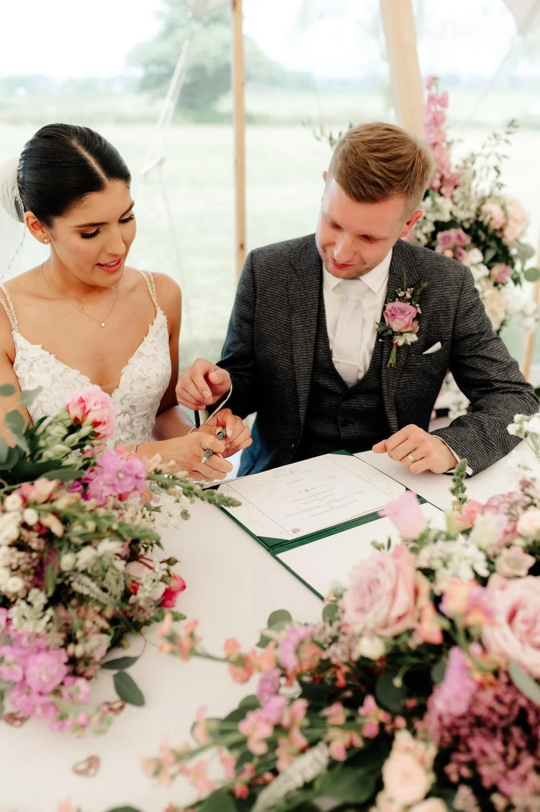 A bride and groom signing their marriage certificate at a wedding ceremony, surrounded by pink and white flowers.