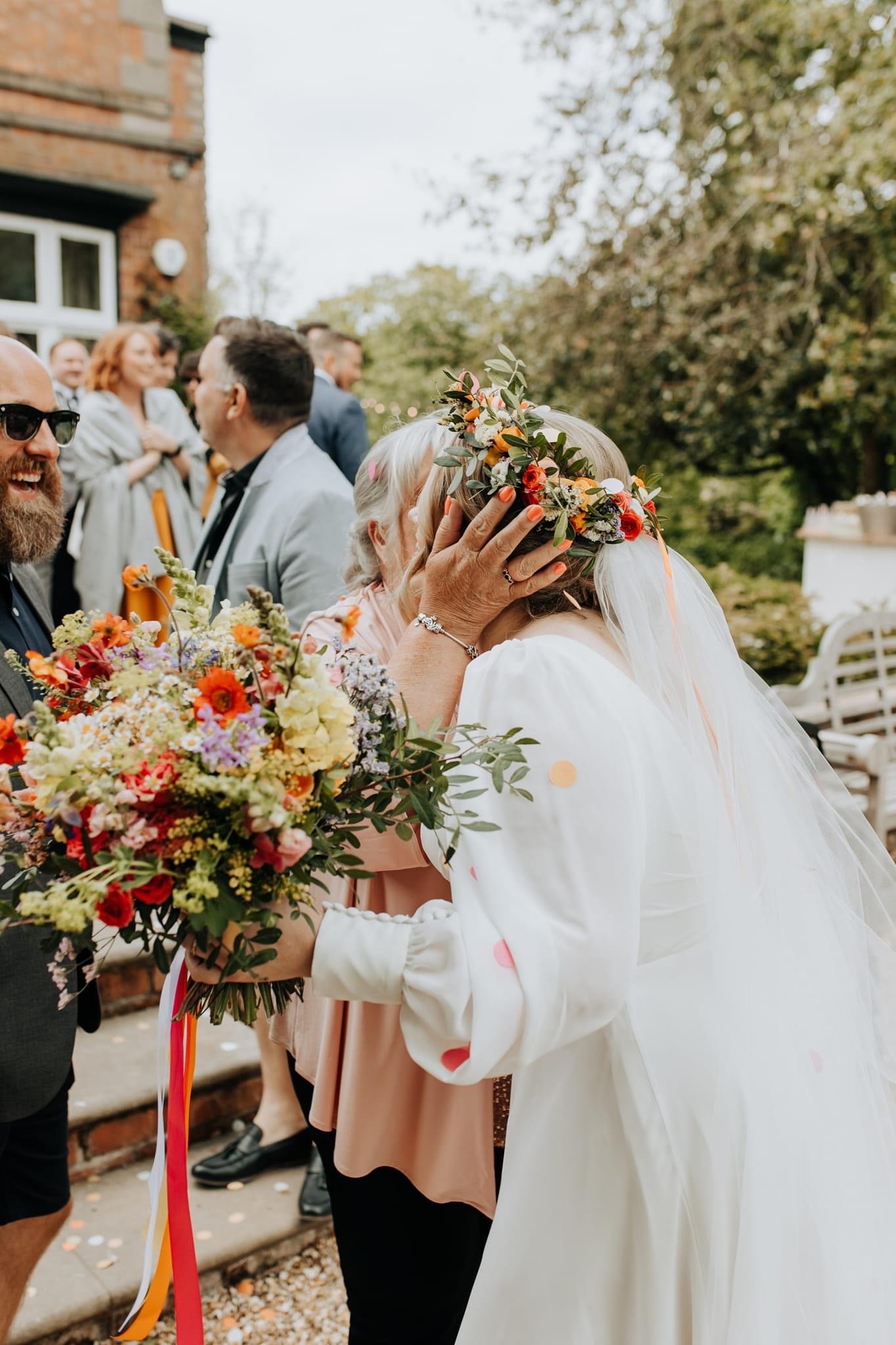 A bride wearing a floral crown is being kissed on the forehead by a woman, possibly her mother, at a wedding reception outdoors. The bride holds a large bouquet of colorful flowers and is dressed in a white gown, with guests and greenery in the backg