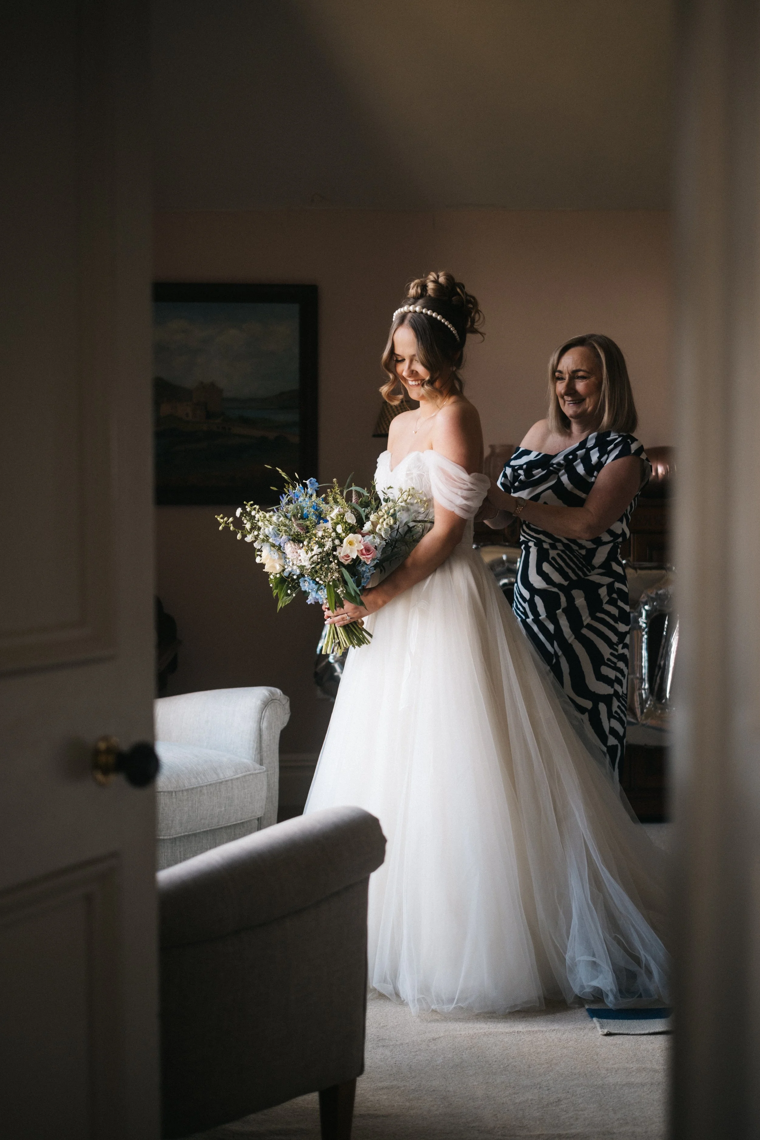 A bride in a white wedding dress holding a bouquet of flowers, smiling, standing in a living room with a woman helping her with her dress or veil, and various furniture items like a gray armchair and a painting on the wall.