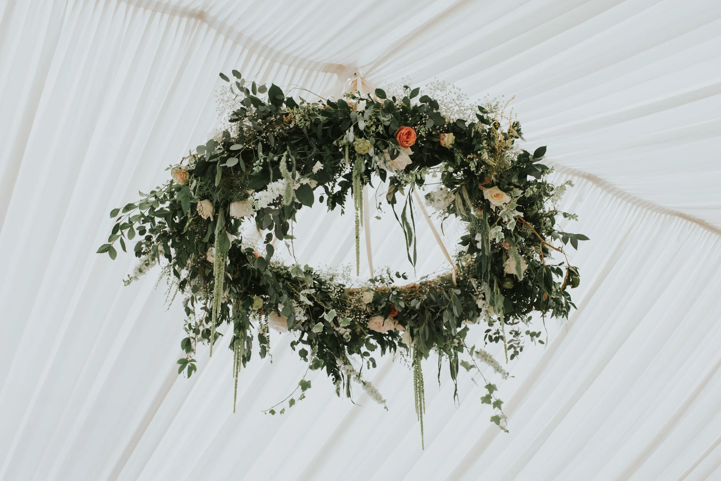A floral arrangement hanging from a ceiling with white drapery, featuring green leaves and various flowers including roses.