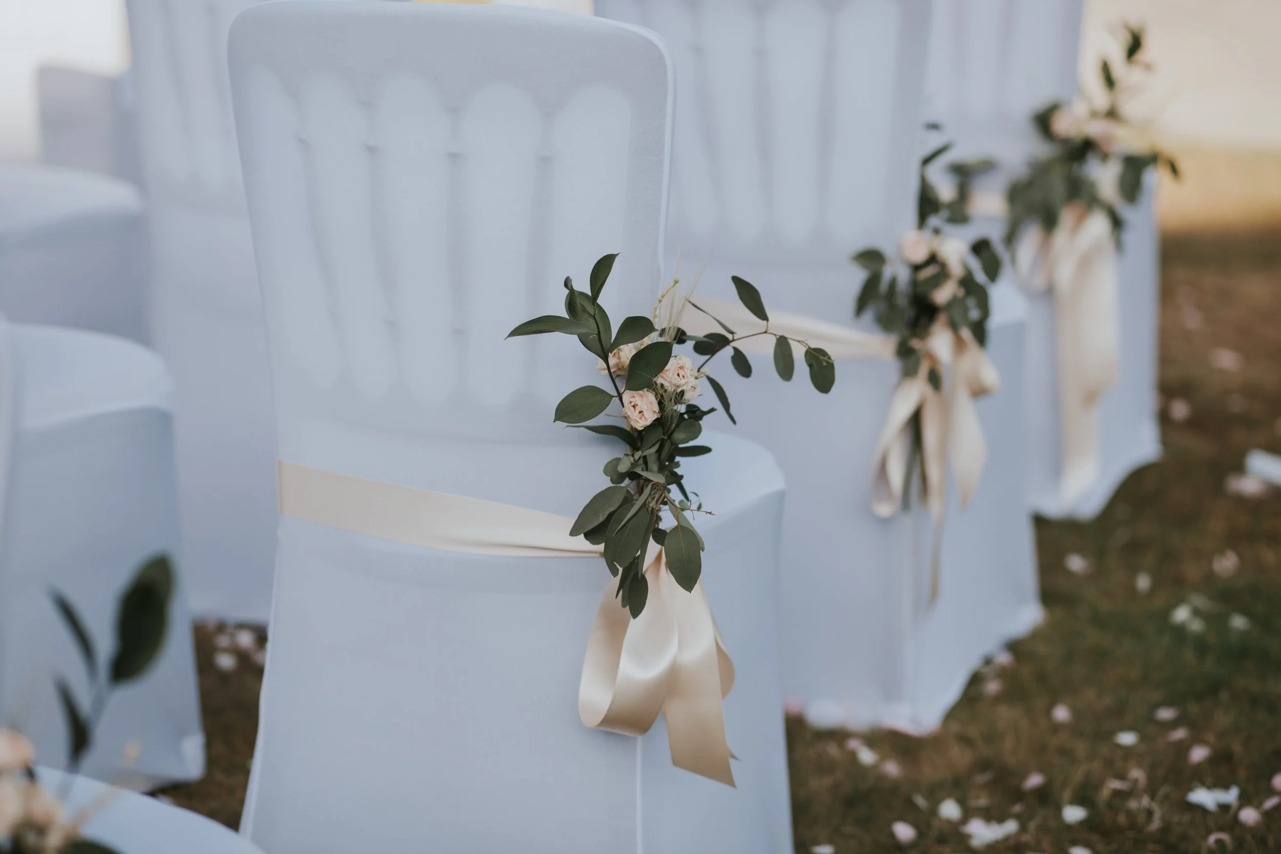 White chairs decorated with small bouquets of pink roses and green leaves tied with beige ribbons, arranged in a row outdoors.