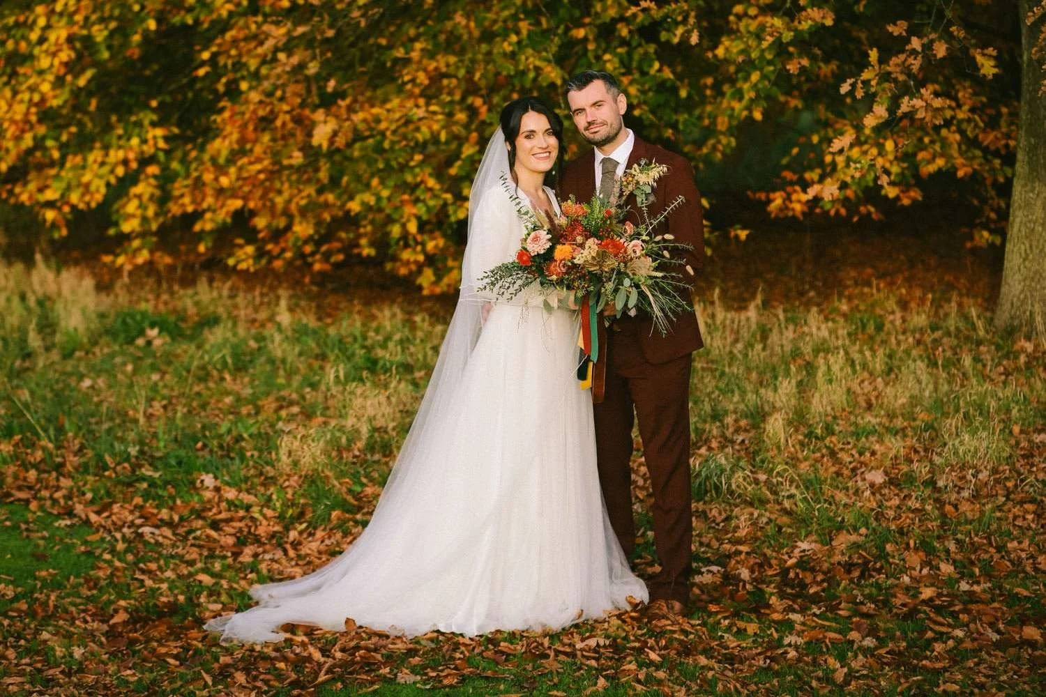 A bride and groom standing together outdoors during fall, with yellow and orange leaves on the trees and ground, the bride wearing a white wedding dress and veil, holding a large bouquet of flowers, and the groom in a dark suit.