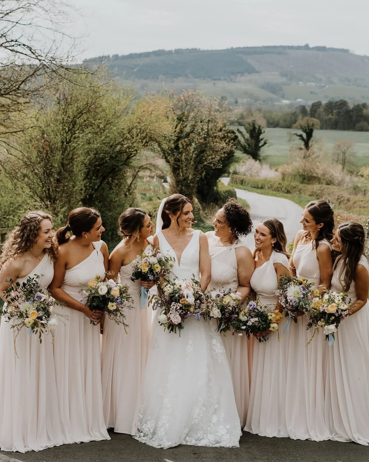 Group of women in wedding dresses holding bouquets, standing outdoors on a rural road with trees and rolling hills in the background.
