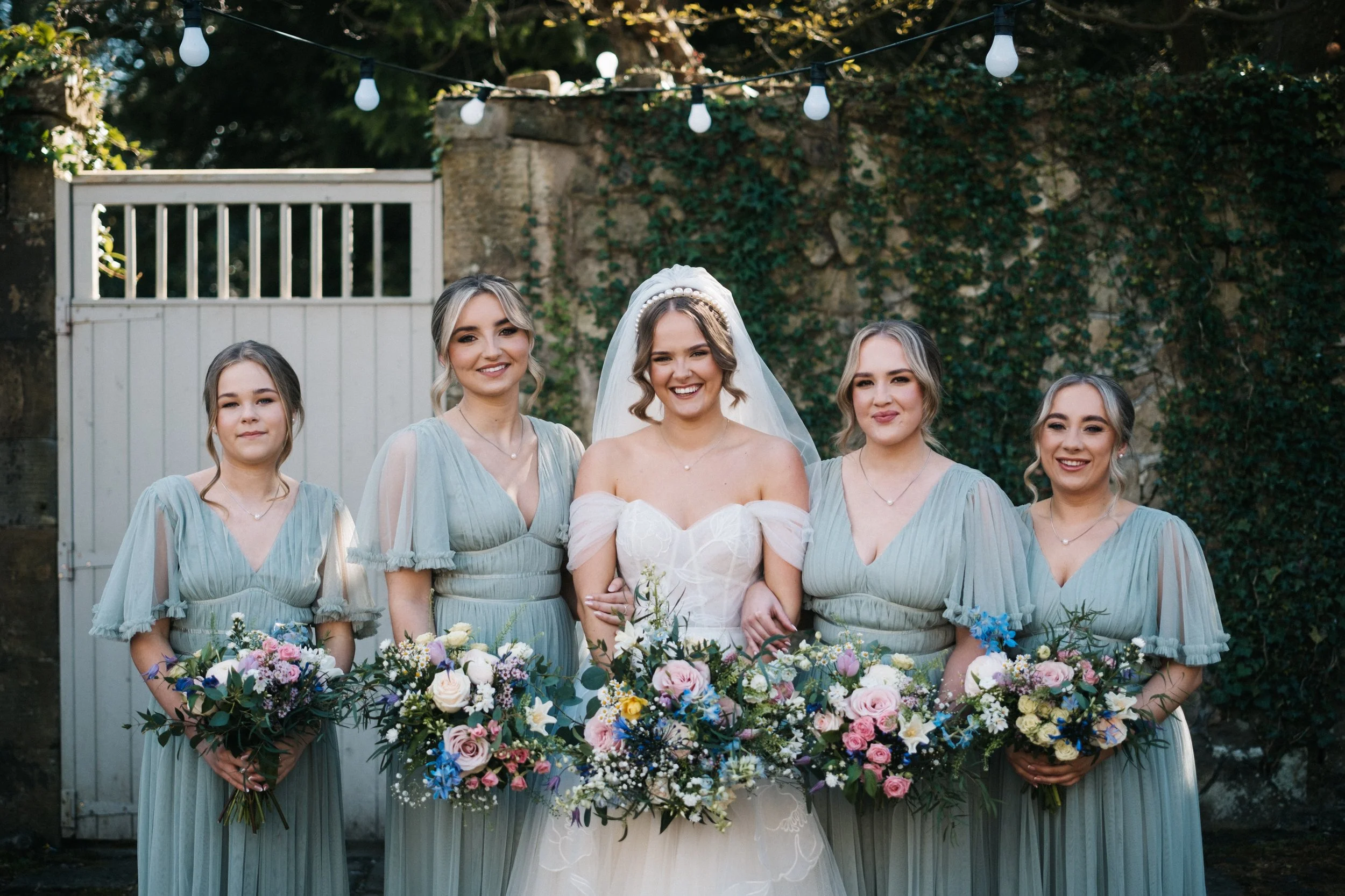 A group of five women, including a bride in a white wedding dress with a veil, and four bridesmaids in matching light blue dresses, standing outdoors in front of a brick wall covered in ivy. They are smiling and holding large bouquets of flowers.