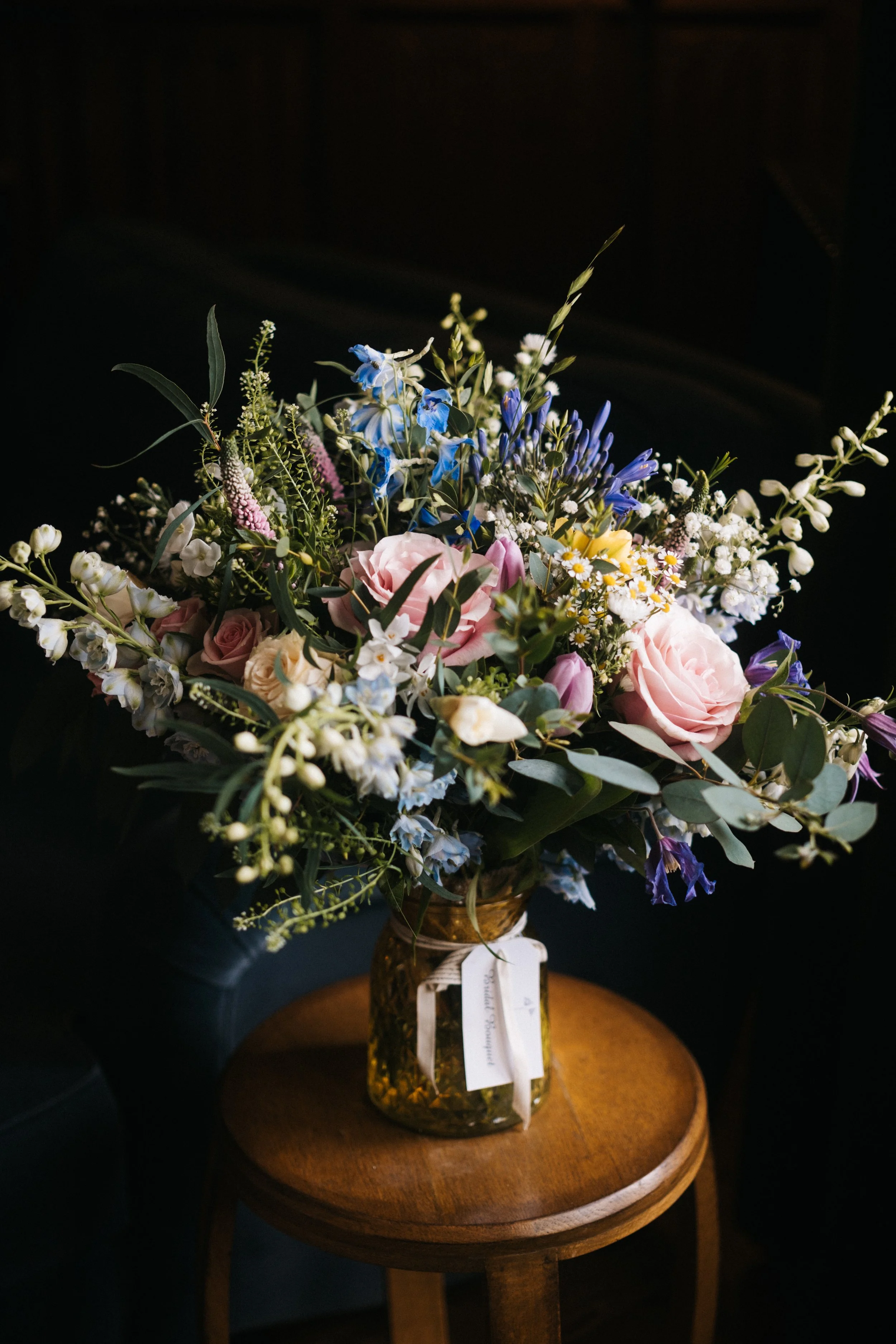 A bouquet of mixed flowers in a vintage amber glass jar on a wooden table, with a black background.