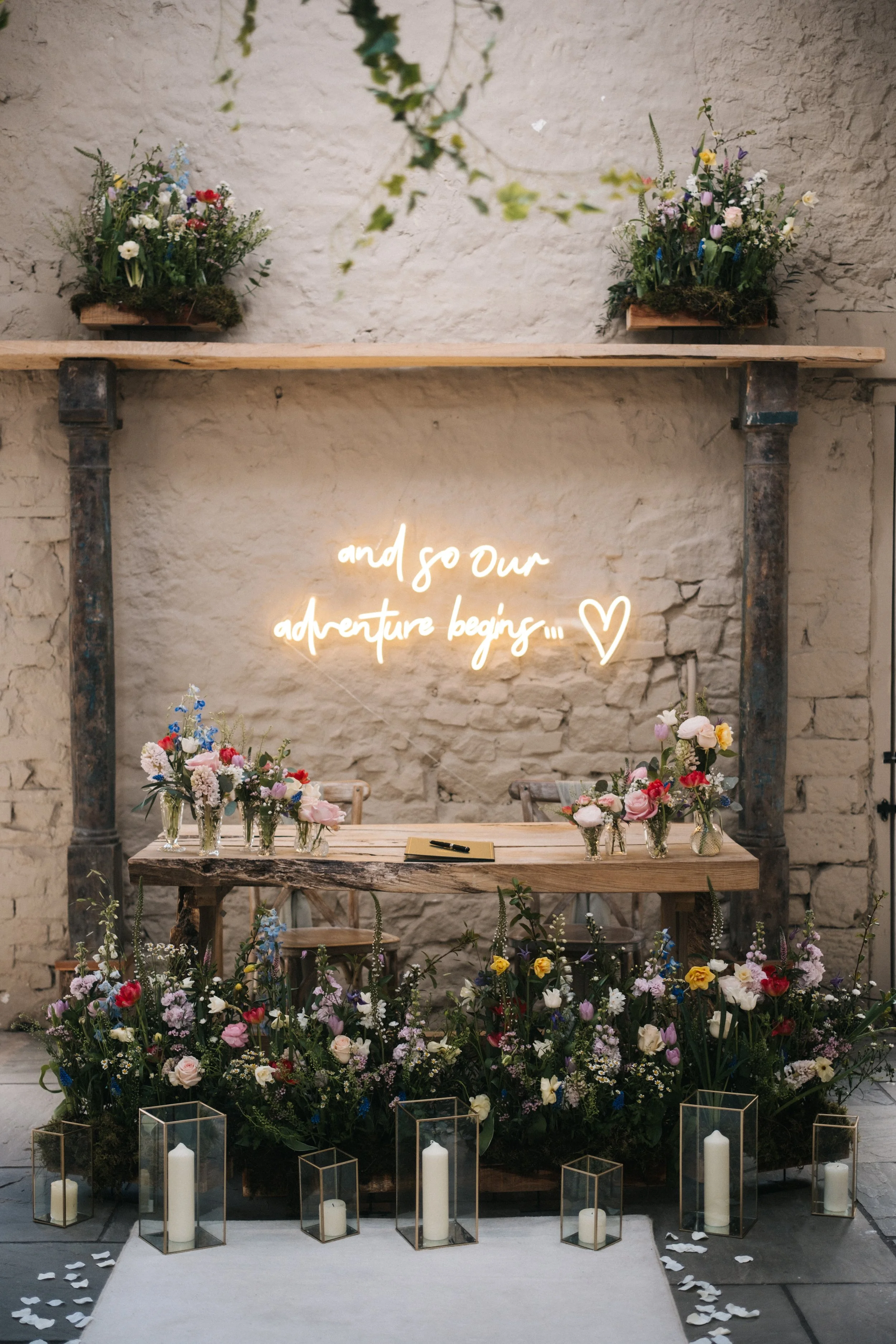 Wedding altar with a rustic wooden table adorned with multiple small vases filled with colorful flowers, surrounded by glass candle holders with white candles, against a textured stone wall. A neon sign with the words 'and so our adventure begins...'