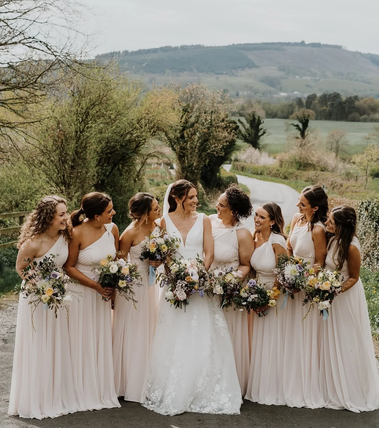 A group of women in wedding dresses standing outdoors in a countryside setting, smiling and holding bouquets of flowers.