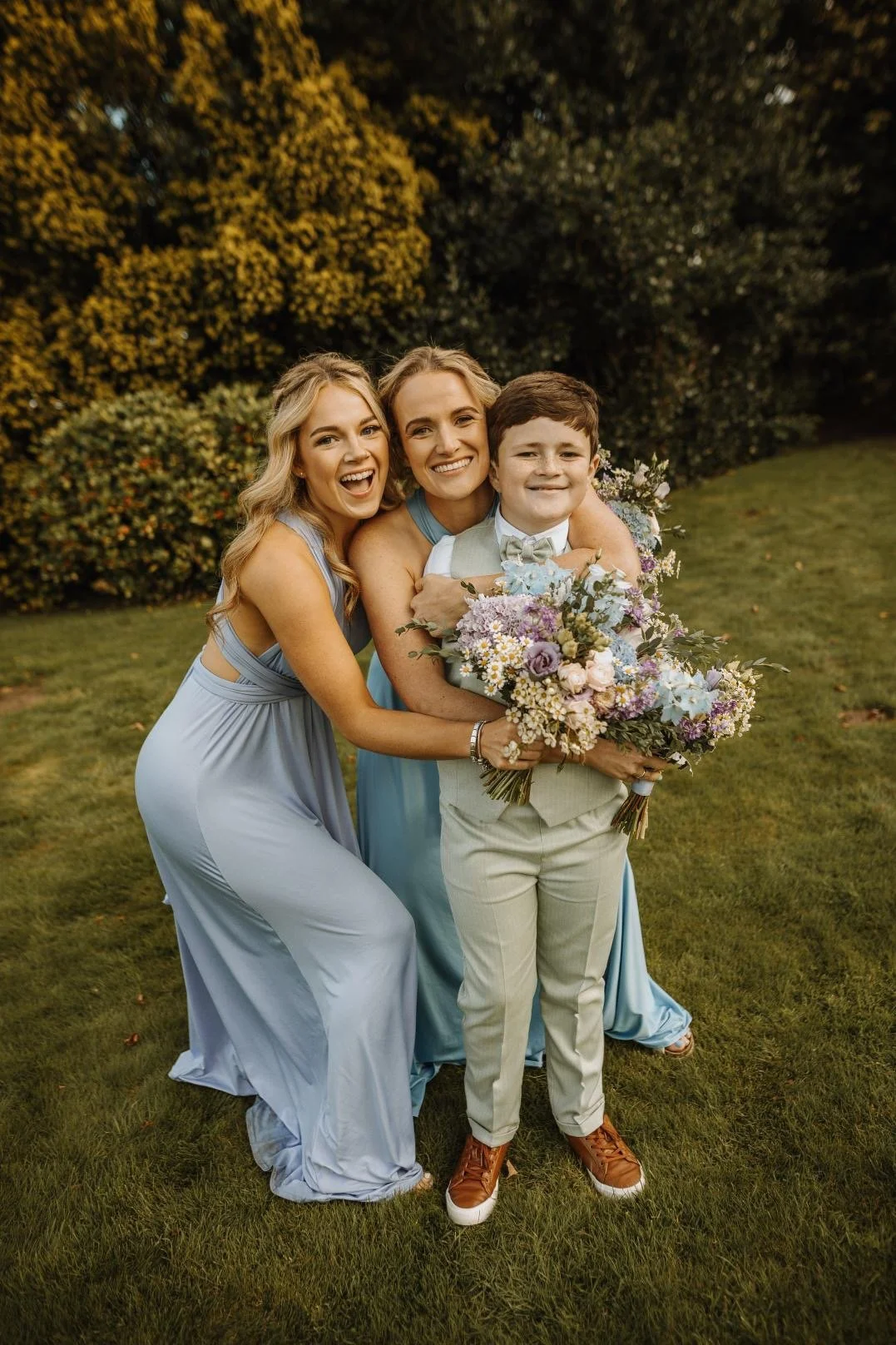 Three people, two women and a boy, smiling and posing together outdoors with trees and grass in the background. One woman and the boy are holding a bouquet of flowers.