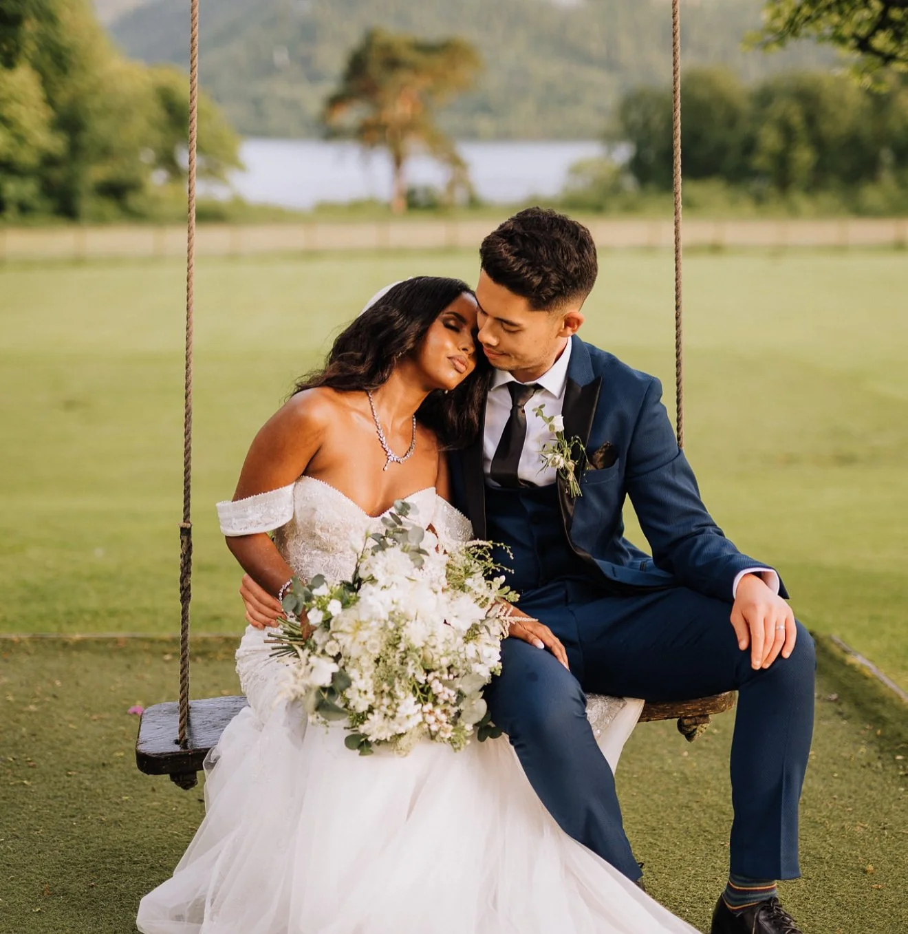 A bride and groom sitting closely on a wooden swing at an outdoor wedding venue, with a lake and trees in the background, the bride in a white off-the-shoulder wedding dress holding a large bouquet of white flowers, and the groom in a navy blue suit 
