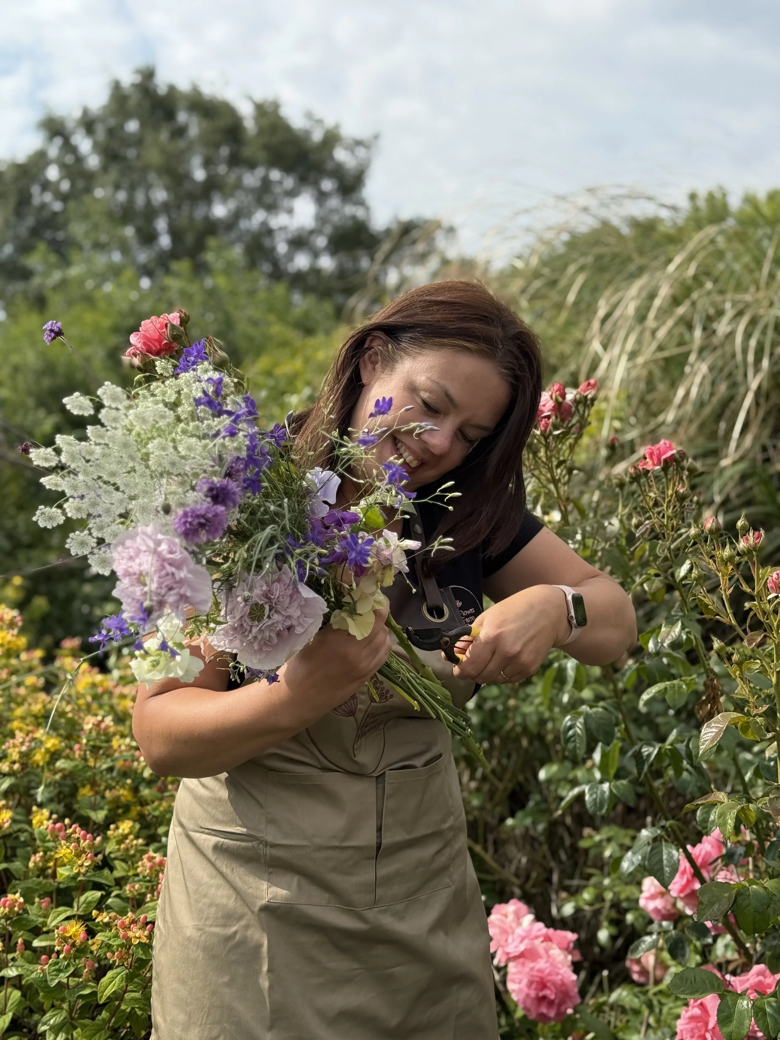 A woman with brown hair smiling while cutting flowers in a garden filled with colorful flowers, including pink, purple, and white blooms, with green foliage and trees in the background.