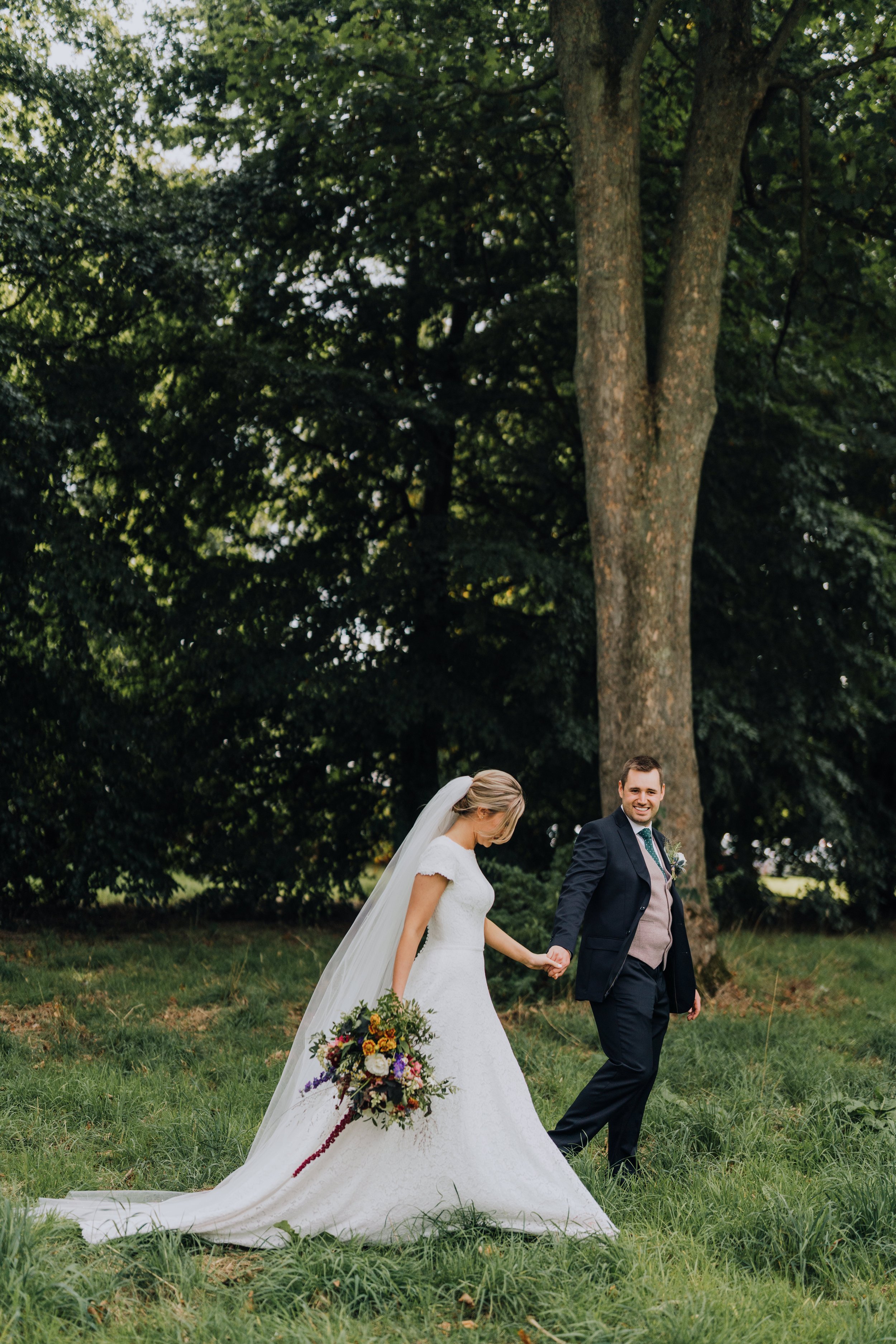 A bride and groom holding hands and walking through a grassy area outdoors near a large tree, with the bride holding a colorful bouquet and wearing a white wedding gown and veil, and the groom in a suit with a boutonniere. They are smiling and lookin
