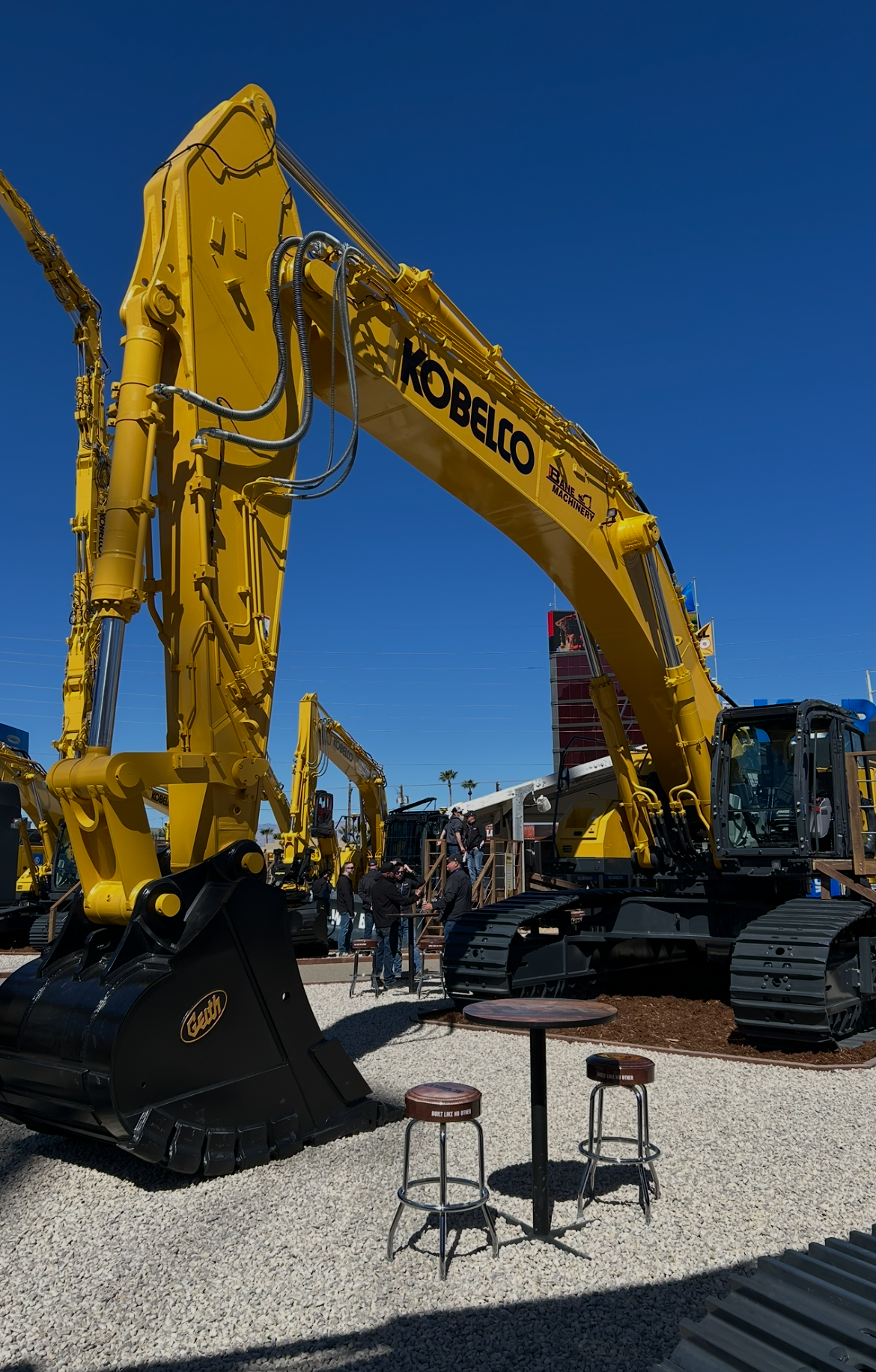 Large yellow Kobelco excavator on display at an outdoor event with people gathered around, and another similar excavator visible in the background under a clear blue sky.