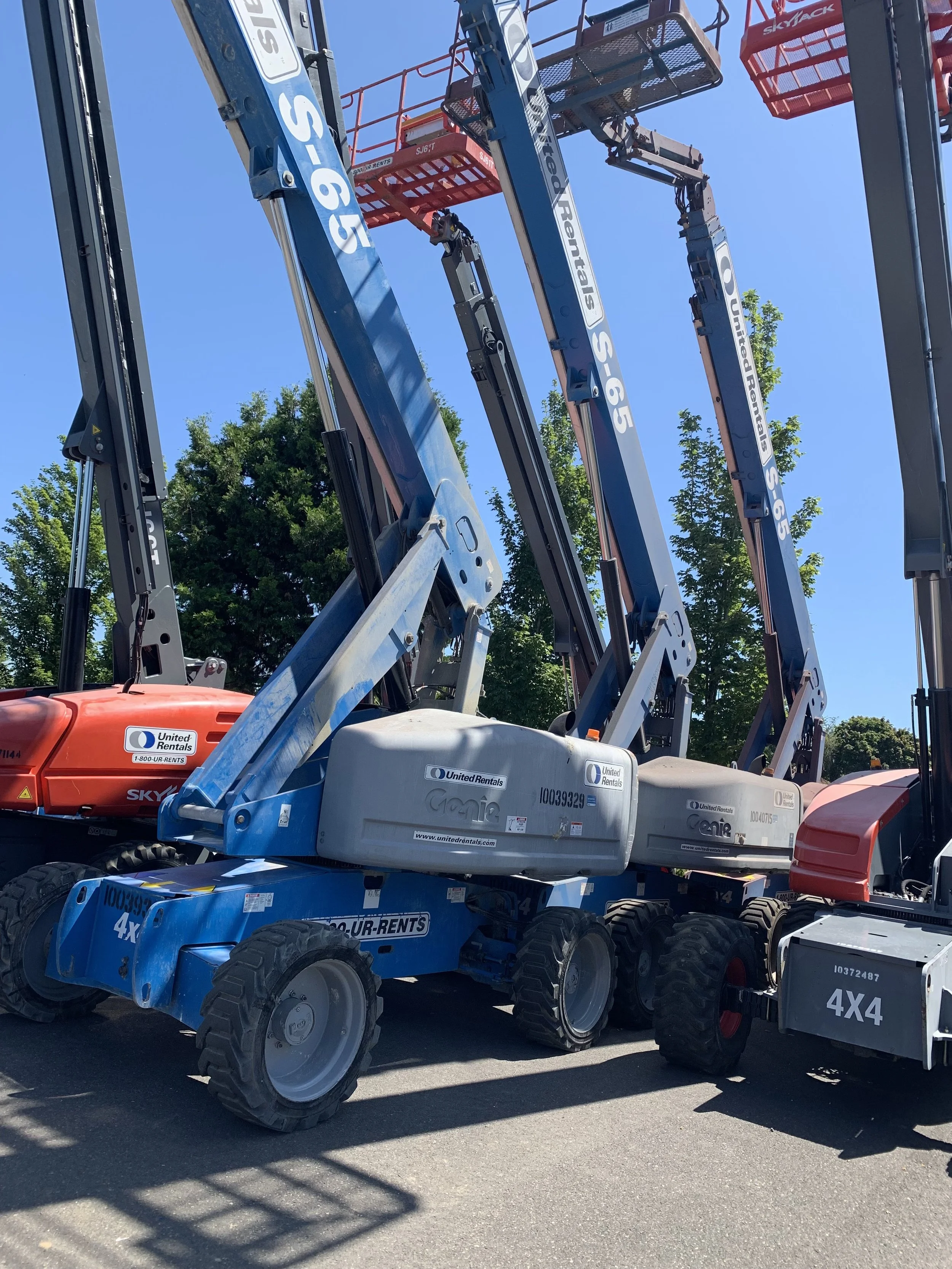 Multiple aerial work platforms with blue, orange, and red arms parked outdoors under a clear blue sky, with green trees visible in the background.