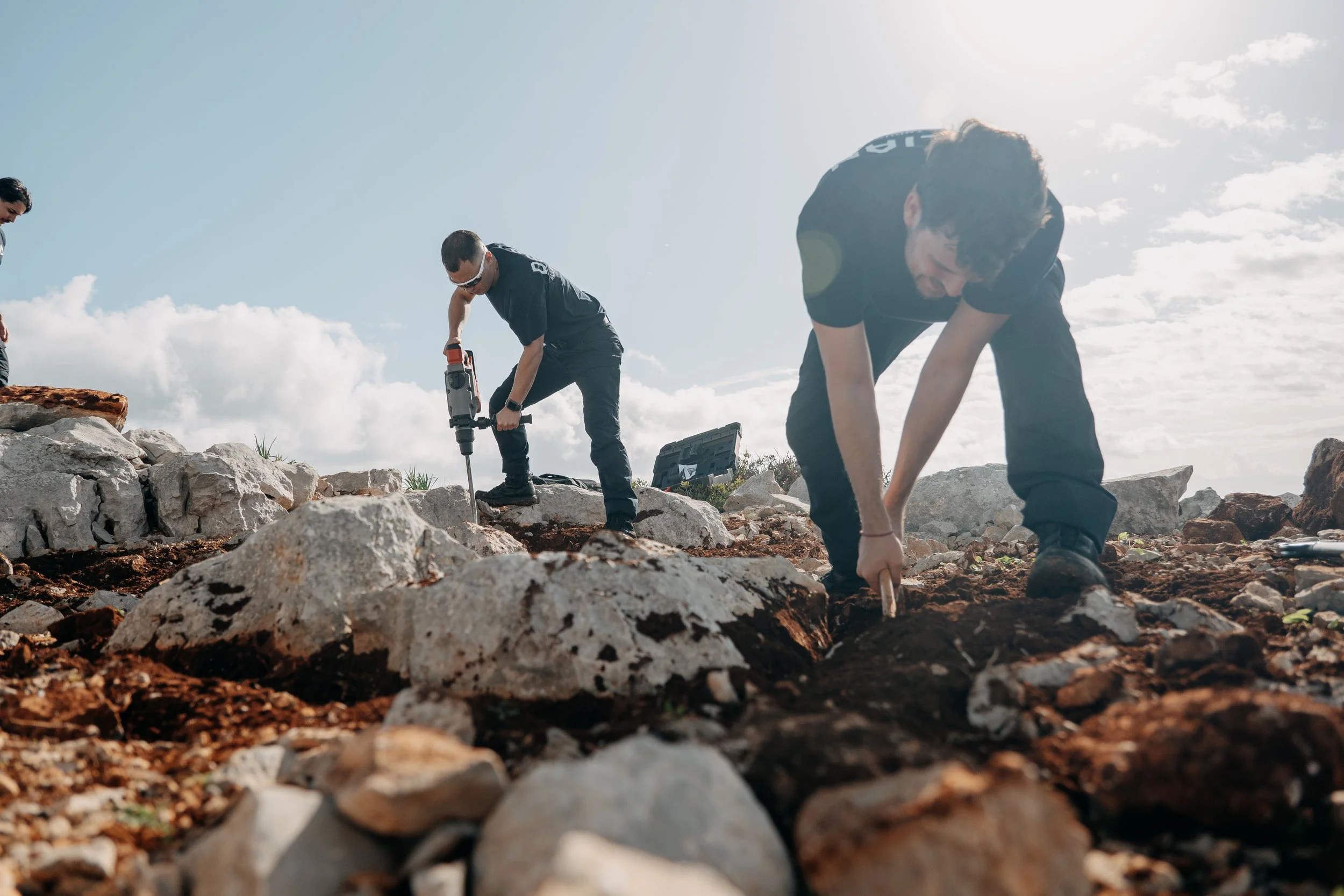 Three people working outdoors, using tools to break rocks on rough terrain under a sunny sky.