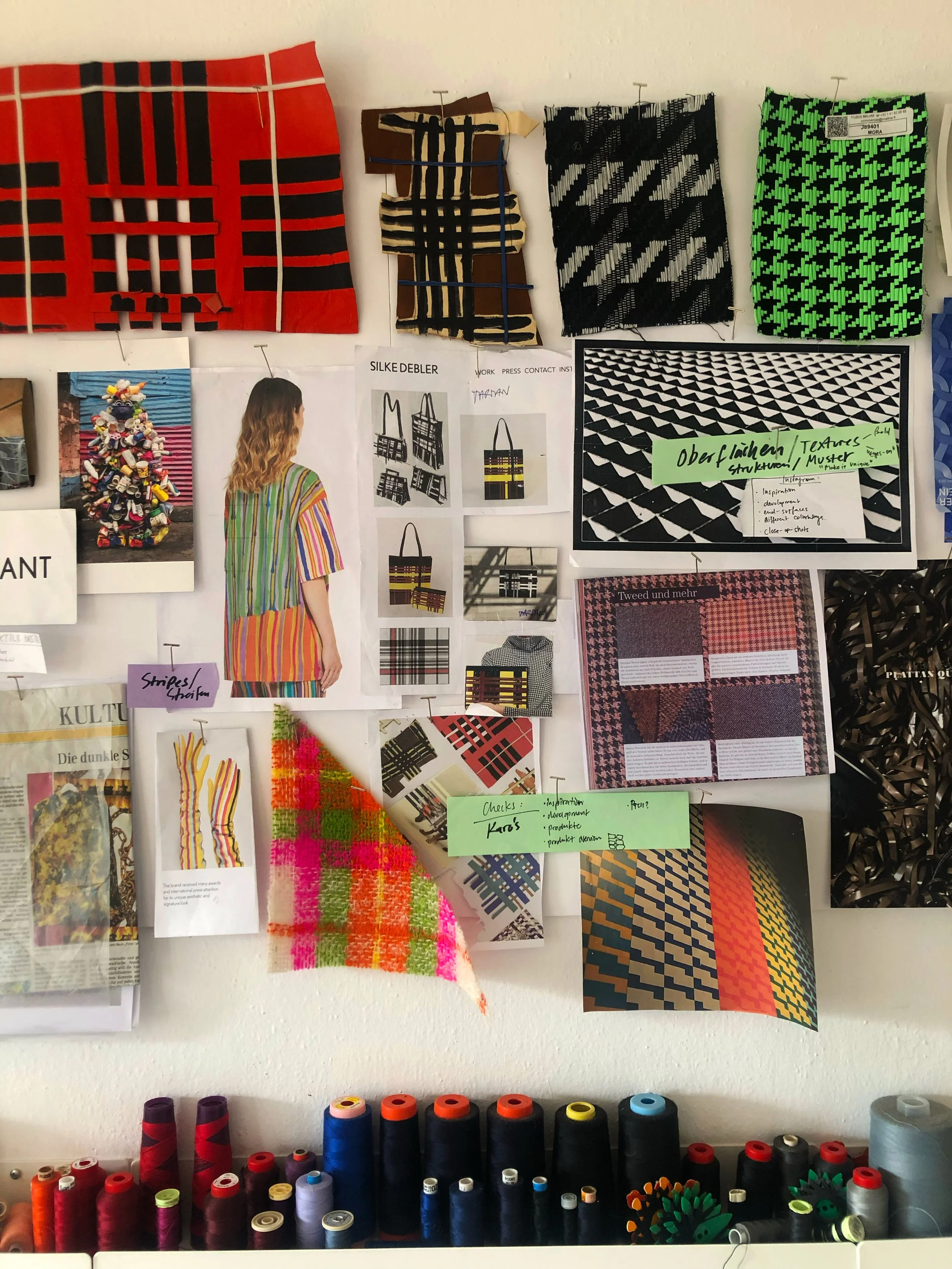 Display of colorful fabrics and textiles with various patterns, sewing and design notes, and spools of thread on a work table.