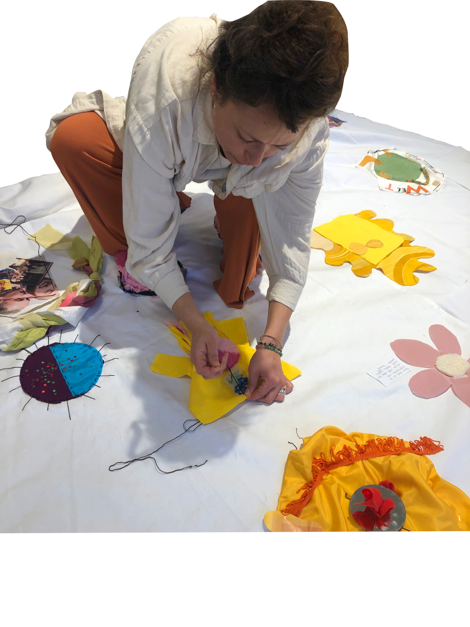 A woman is kneeling on a white surface, working on colorful fabric art pieces, with materials and completed fabric designs around her.