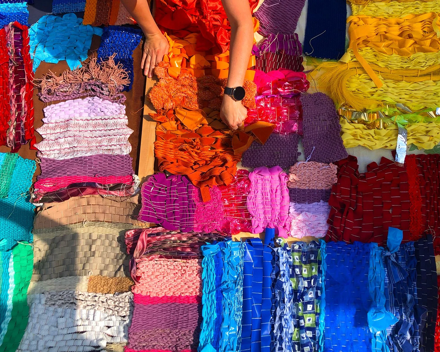 A person arranging colorful woven ribbons and fabric strips, with various bright and pastel paper and fabric crafts on a table.