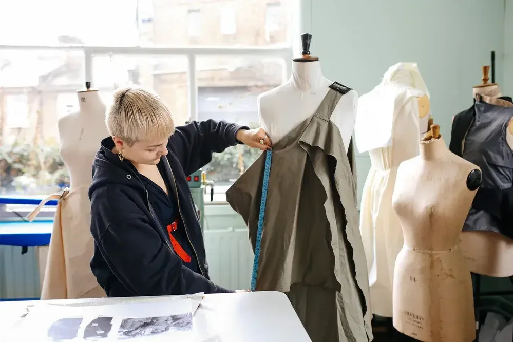 A young person measuring a dress on a dress form in a fashion design studio with multiple dress forms and fabric sketches.