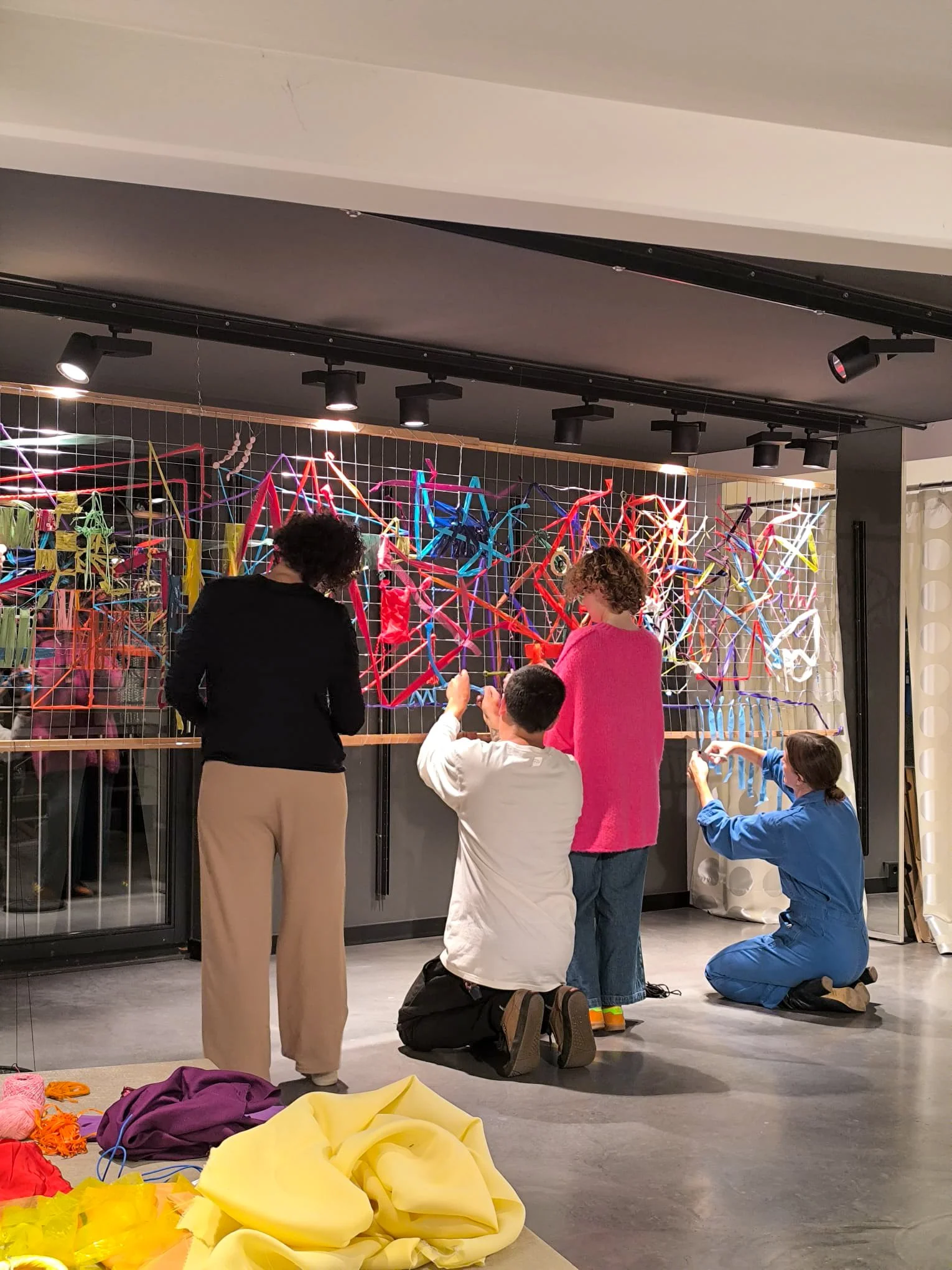 People working on a colorful art installation made of various fabrics and materials behind a wire grid.