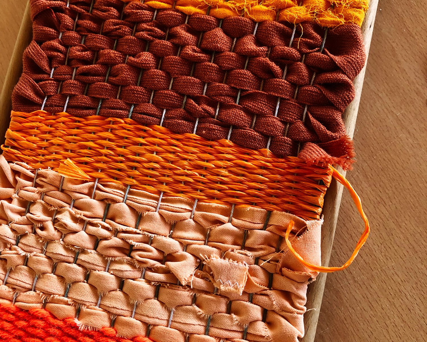 Close-up of three woven textile sample swatches, each with a different weaving technique and color: dark red, orange, and pale pink, laid out on a wooden surface.