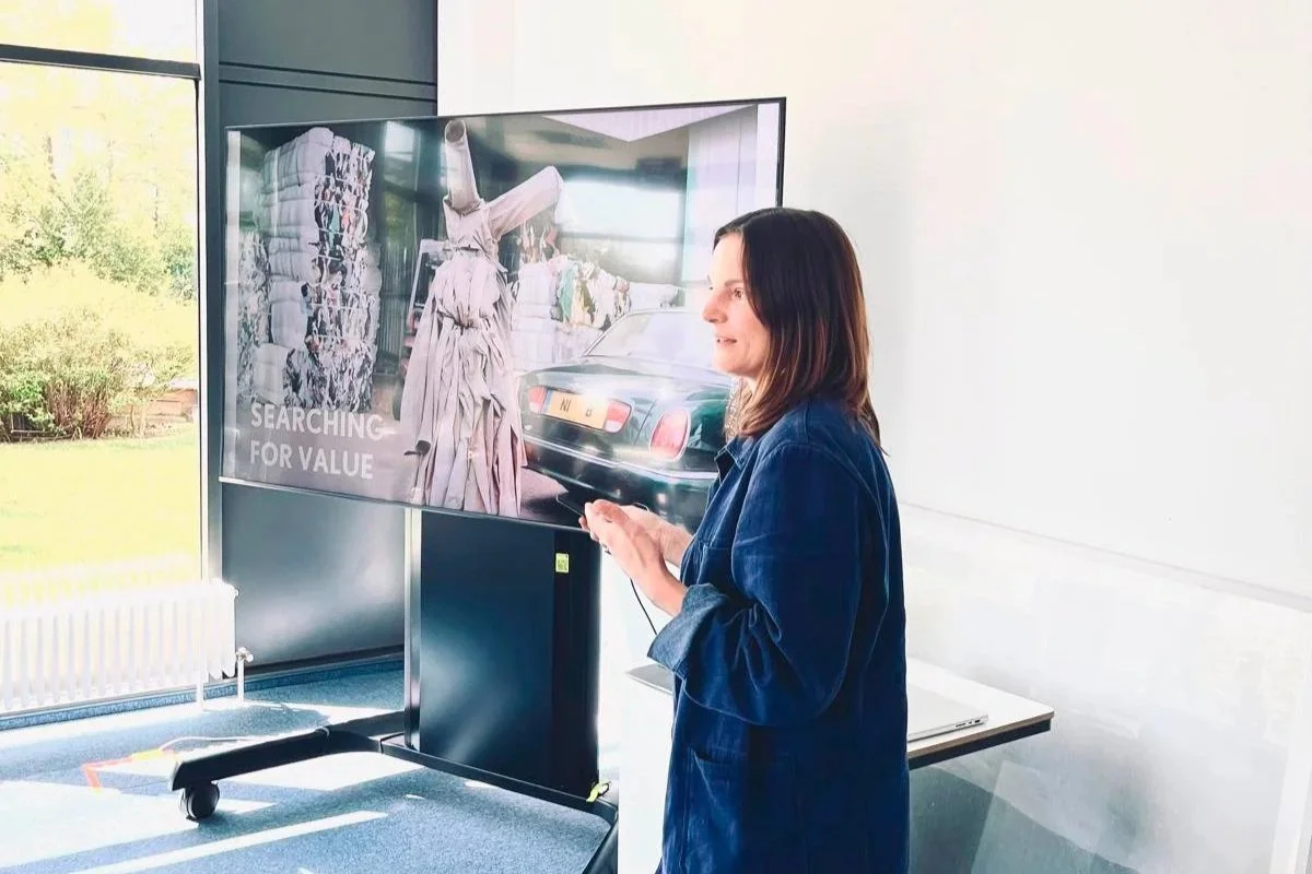 Woman standing in front of a large digital screen, looking at it and using her phone, in a bright room with large windows and a view of trees outside.