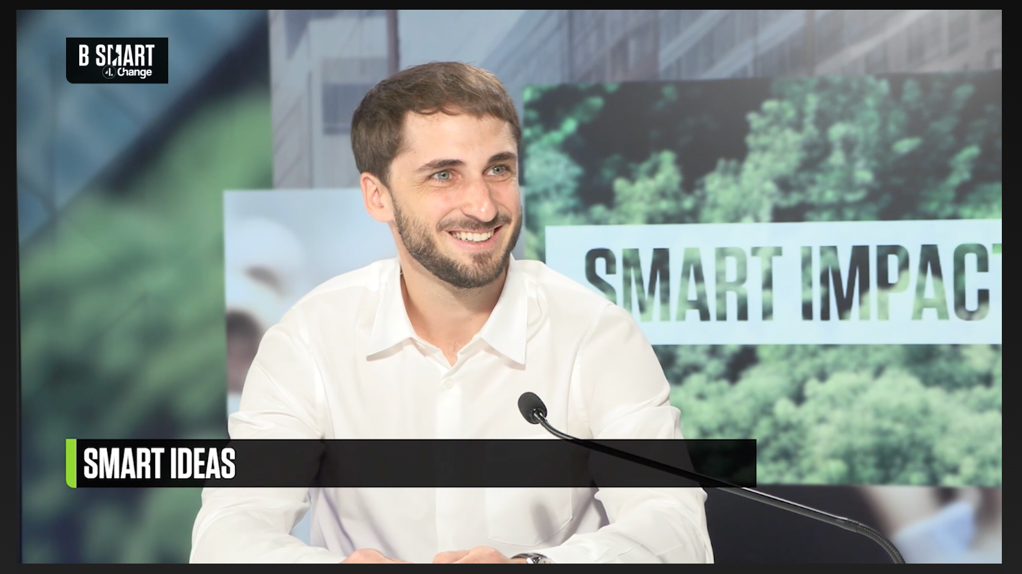 Un homme souriant lors d'une interview ou d'une conférence, avec un micro devant lui. En arrière-plan, un panneau avec le texte "SMART IMPACT" et une scène de nature.