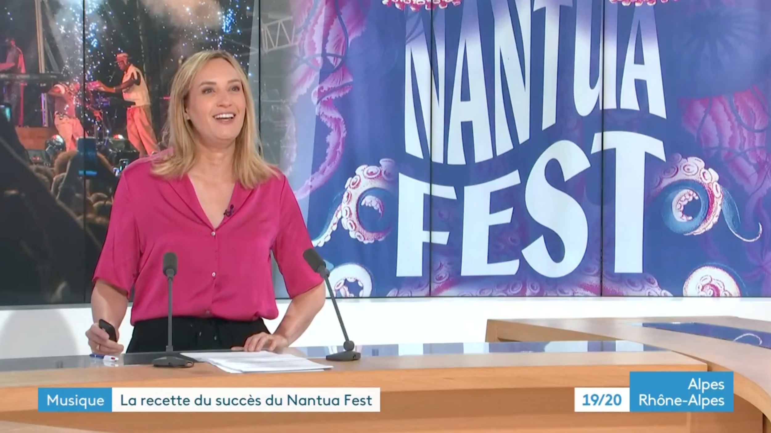 Une femme souriante, portant une chemise rose, parle derrière un pupitre avec deux microphones, lors d'une émission télévisée sur le festival Nantua Fest, en Rhône-Alpes. En arrière-plan, une grande affiche colorée du festival avec des éléments de décoration et le nom du festival.