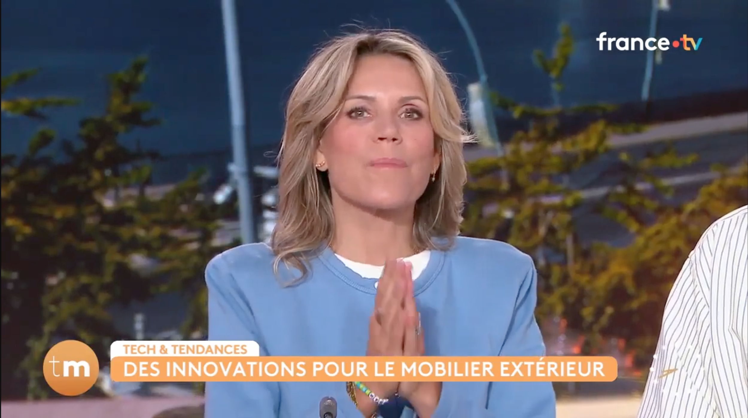 Une femme à cheveux blonds, portant une chemise bleue, participe à une émission télévisée sur France.tv, discutant devant un décor extérieur avec des arbres et un ciel nocturne, avec un bandeau inférieur indiquant des innovations pour le mobilier extérieur.