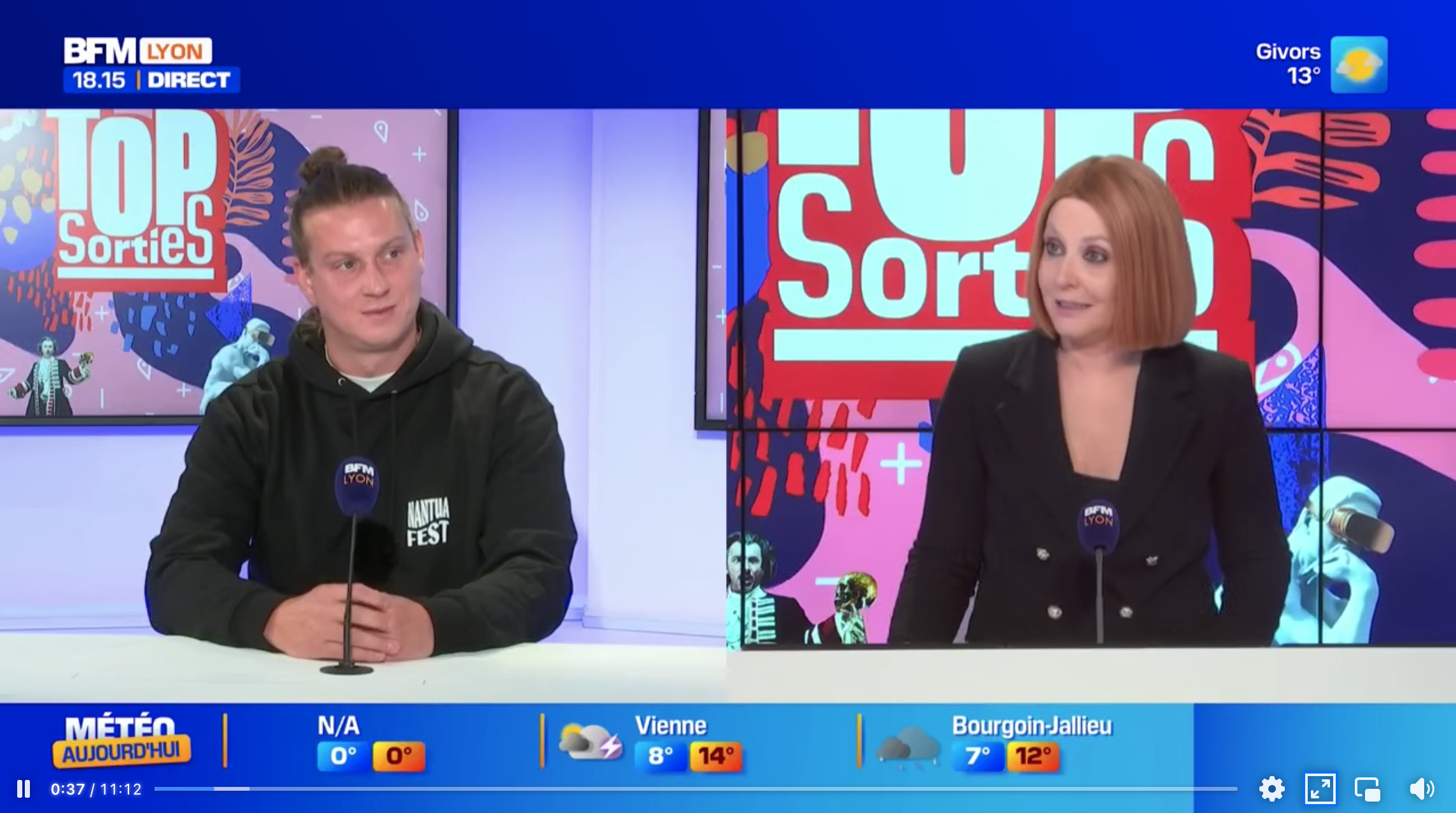 Deux présentateurs de l'émission 'Top Sorties' de BFM Lyon, un homme avec des cheveux attachés en queue de cheval, portant un sweat noir, et une femme avec des cheveux roux, portant un blazer noir, assis derrière des microphones sur un plateau de télévision coloré.