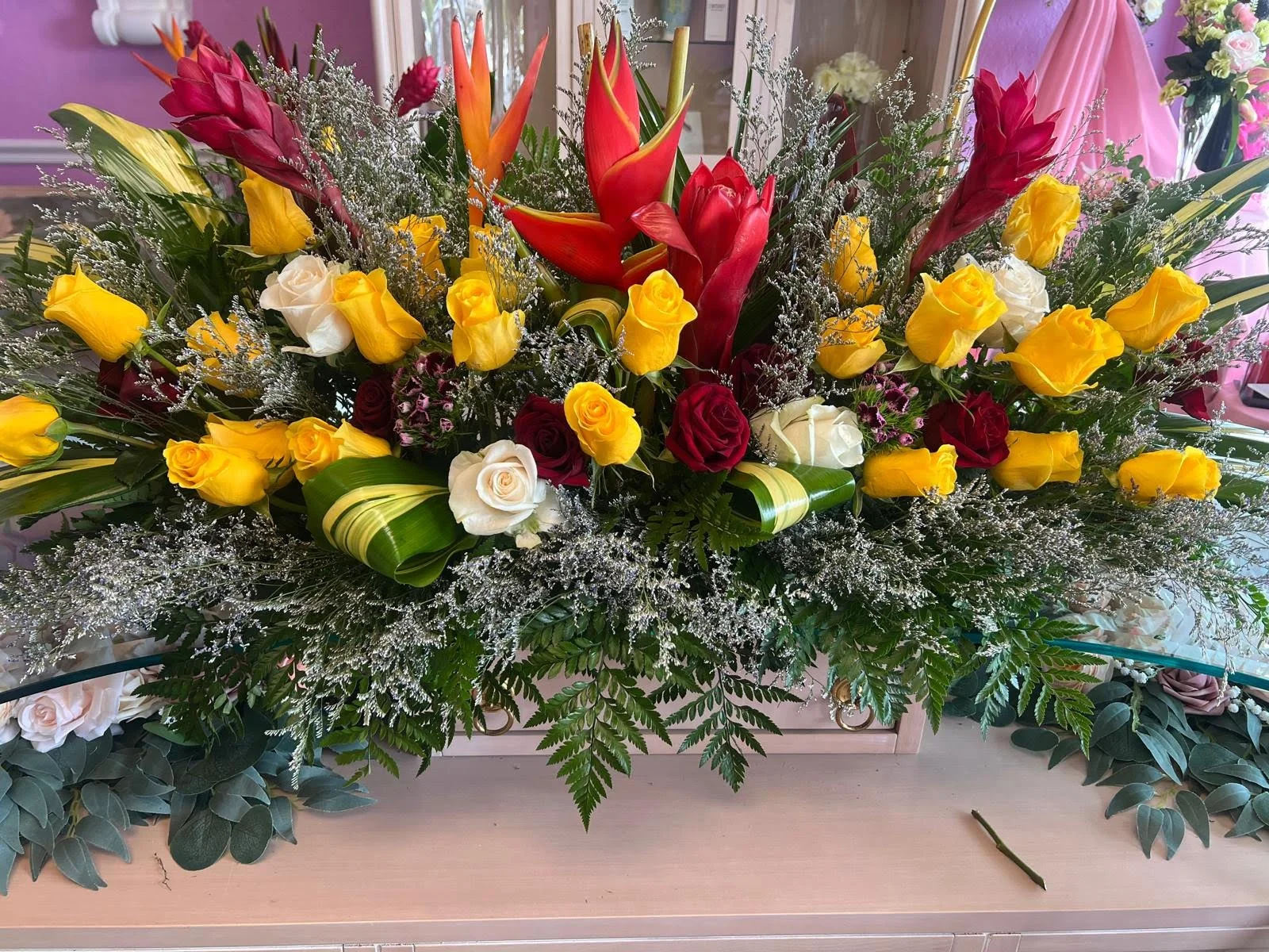 A large floral arrangement featuring yellow, white, and red roses, orange and red tropical flowers, surrounded by green leaves and delicate white filler flowers.