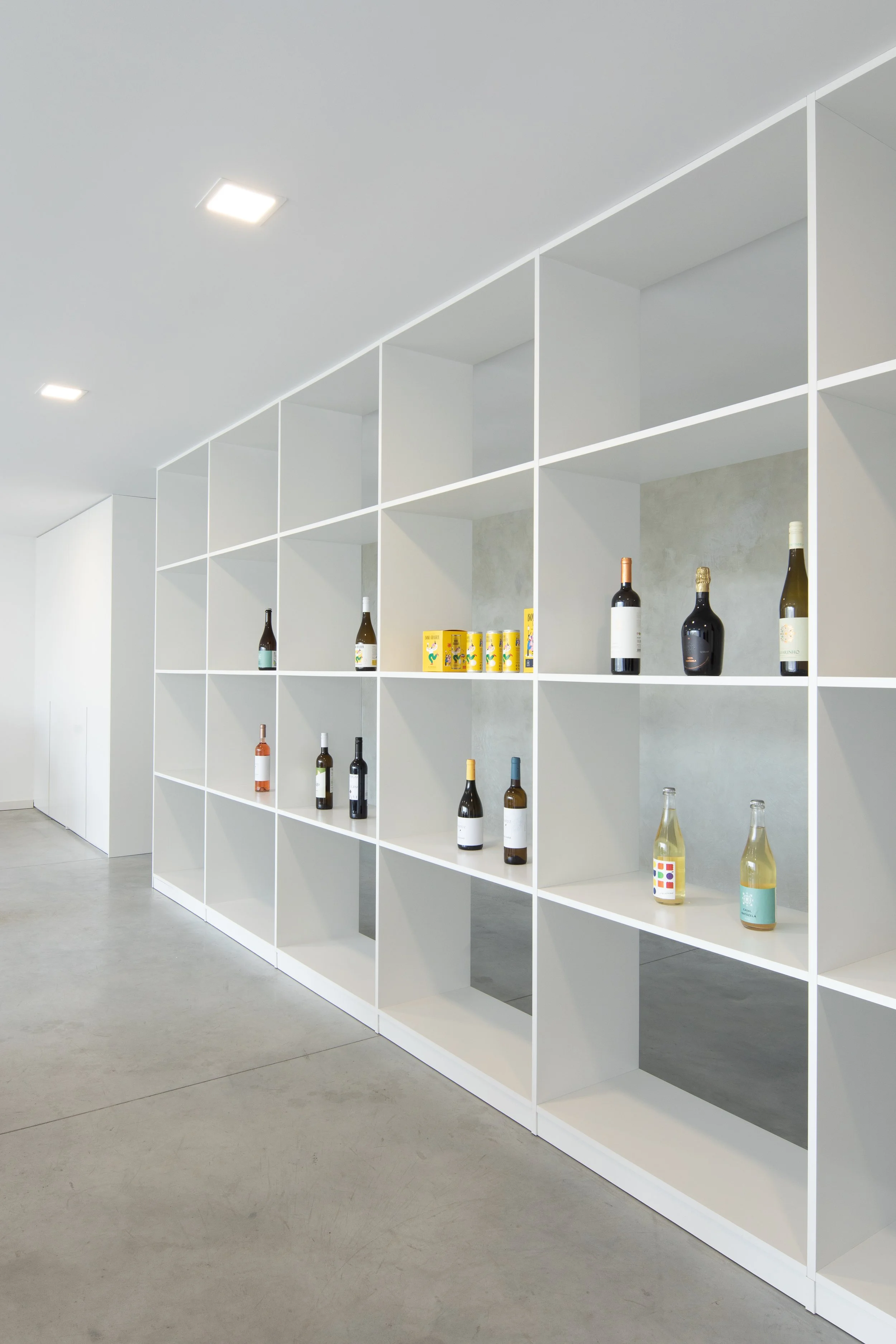 Estante branca com várias garrafas de vinho e algumas latas de alimentos em uma parede de fundo cinza