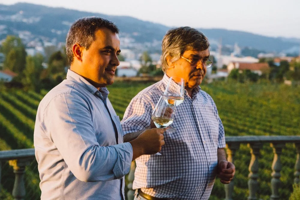 Homens em uma vinícola durante o pôr do sol, um deles segurando uma taça de vinho