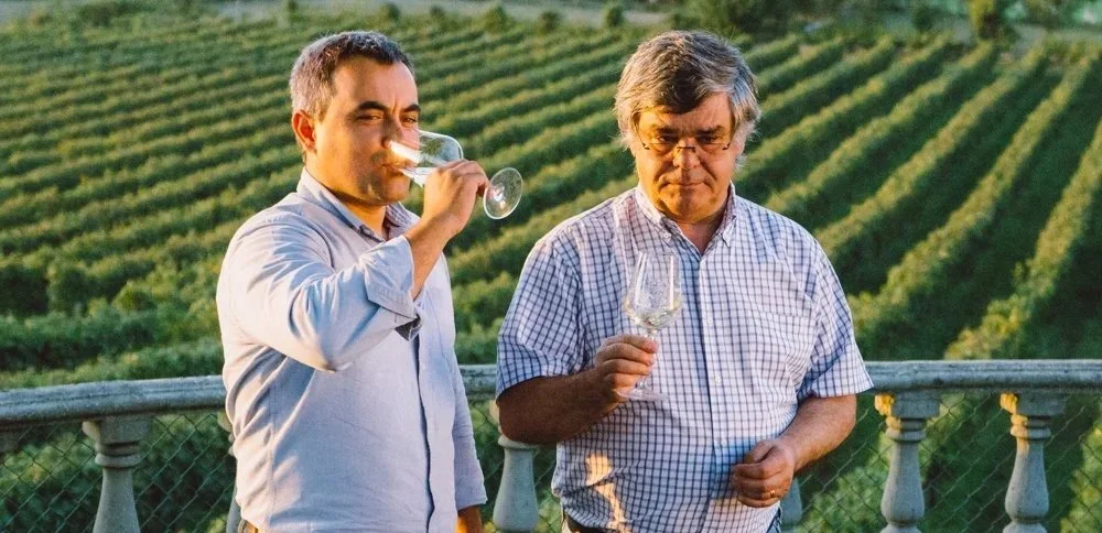 Dois homens degustando vinho em um local ao ar livre com vinhedos ao fundo.