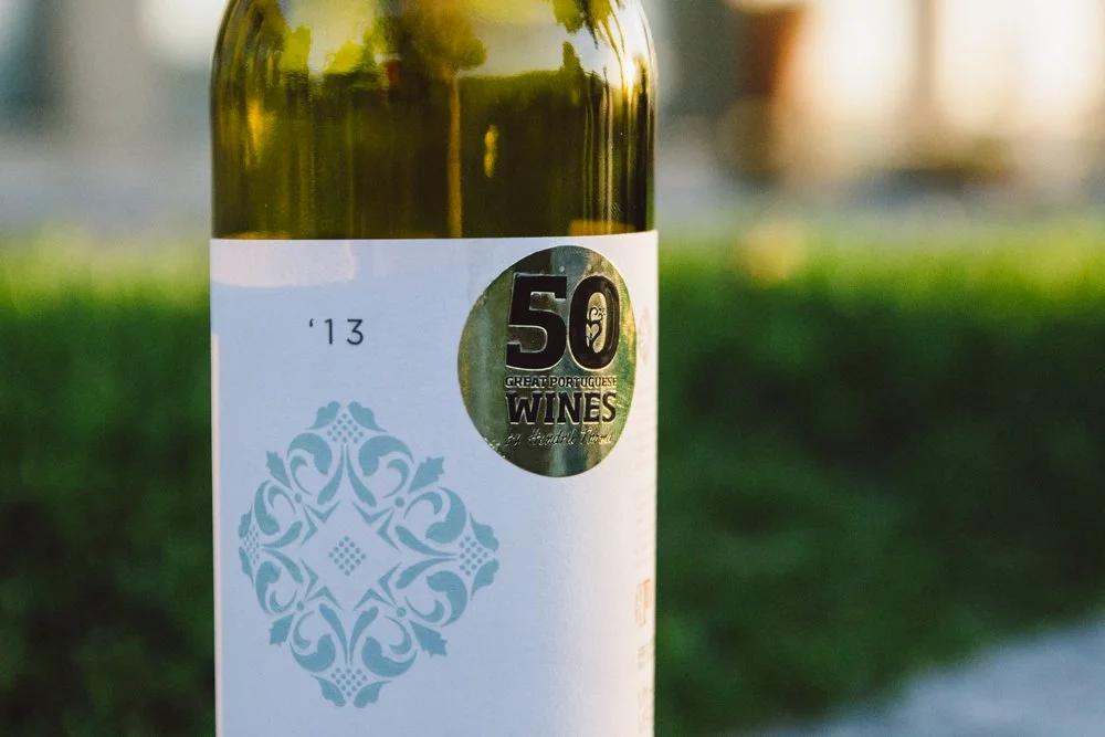 Garrafa de vinho com rótulo branco e selo comemorativo de 50 anos de vinícolas portuguesas, marcada com o ano 2013, ao ar livre em um fundo desfocado de gramado verde.