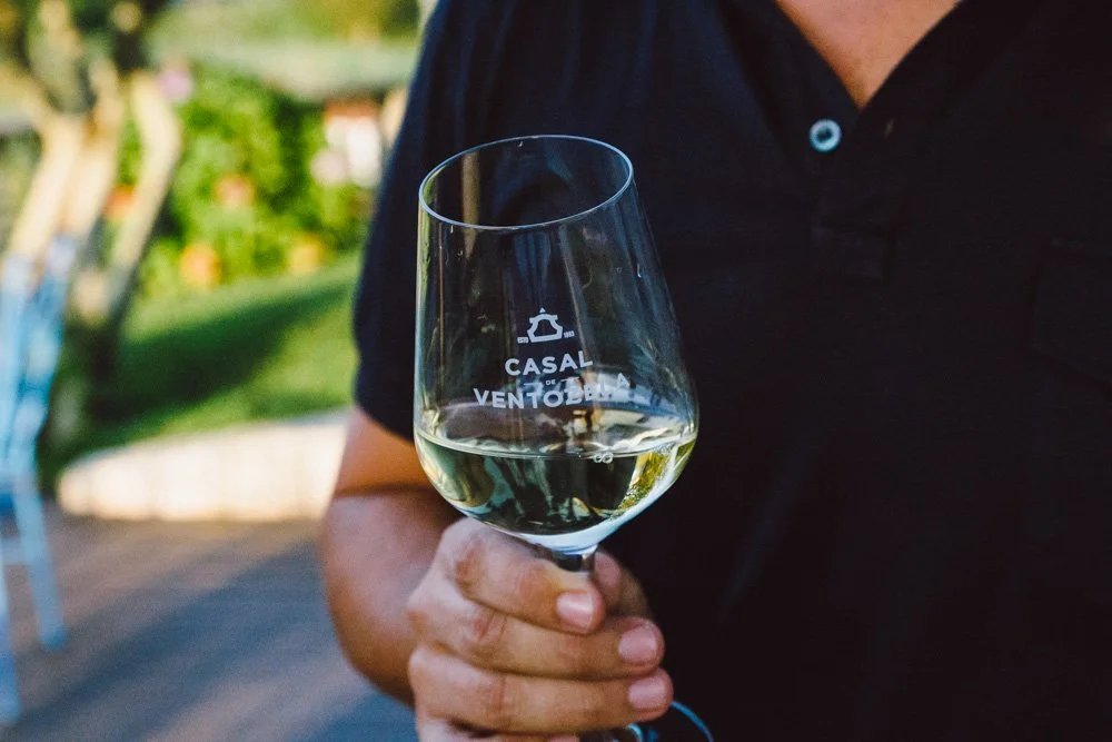 Pessoa segurando taça de vinho branco em um ambiente ao ar livre, com fundo de árvores e jardim.