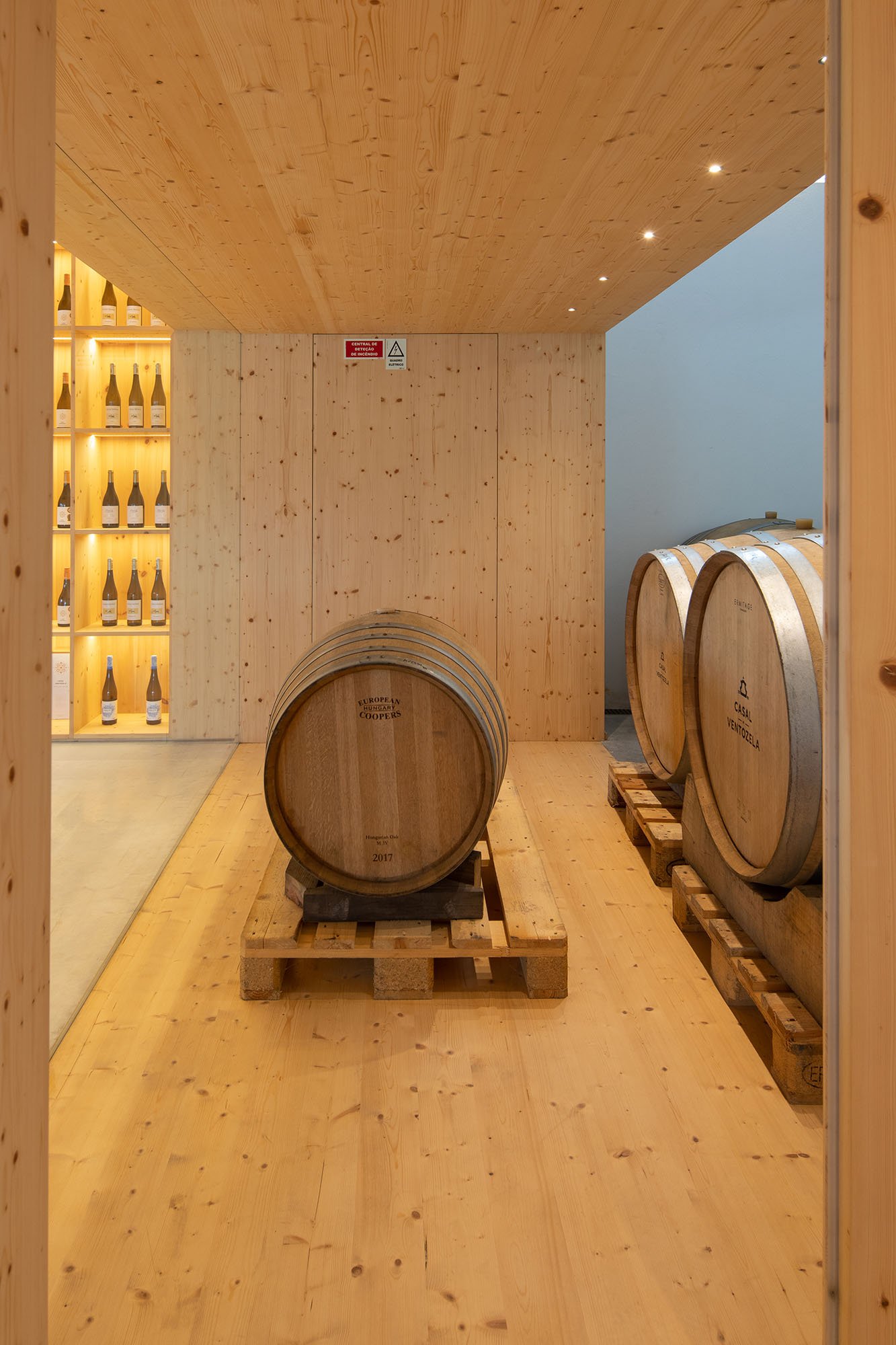Sala de barris de vinho de madeira com garrafas em uma prateleira ao lado.