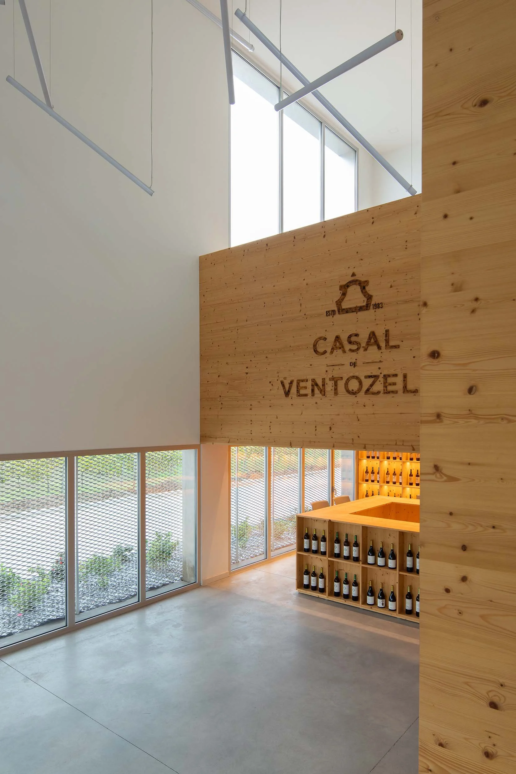 Interior moderno de uma loja de vinhos com paredes de madeira, balcão de degustação e prateleiras com garrafas de vinho, grandes janelas de vidro trazendo luz natural.