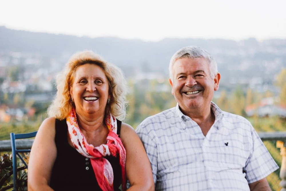 Um casal sorridente, uma mulher de cabelo cacheado loiro e um homem de cabelo grisalho, sentados ao ar livre com vista para uma cidade ao fundo.