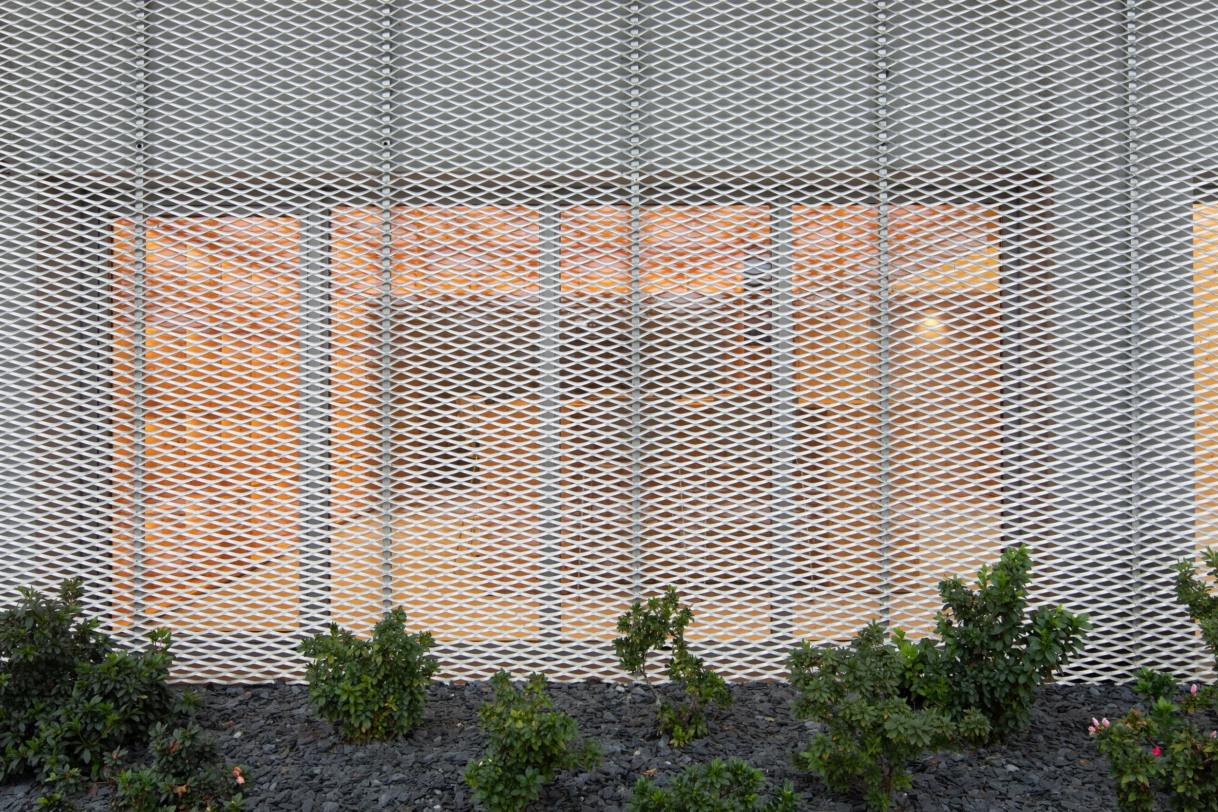 Fachada de um edifício com uma grade de proteção branca na frente, mostrando uma varanda com teto de vidro e móveis ao fundo, plantas verdes na frente, piso de pedras pretas.