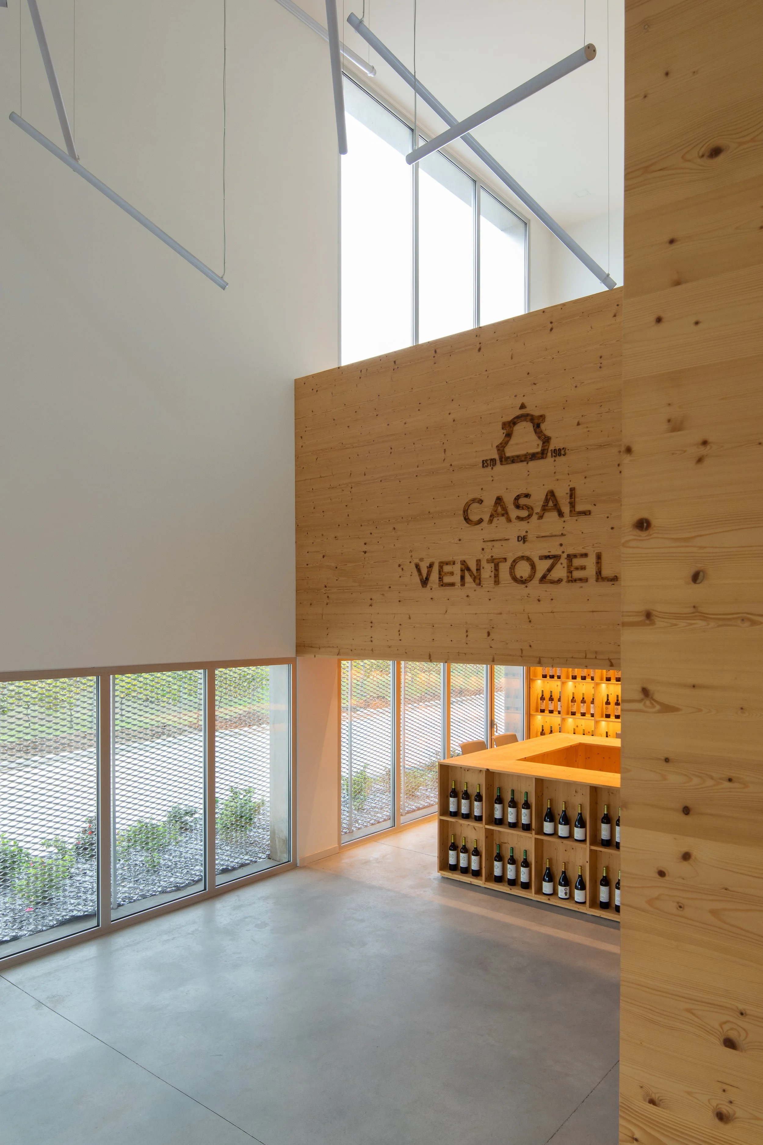 Interior de uma vinícola moderna, com paredes de vidro grandes, prateleiras de madeira com garrafas de vinho e decoração minimalista.