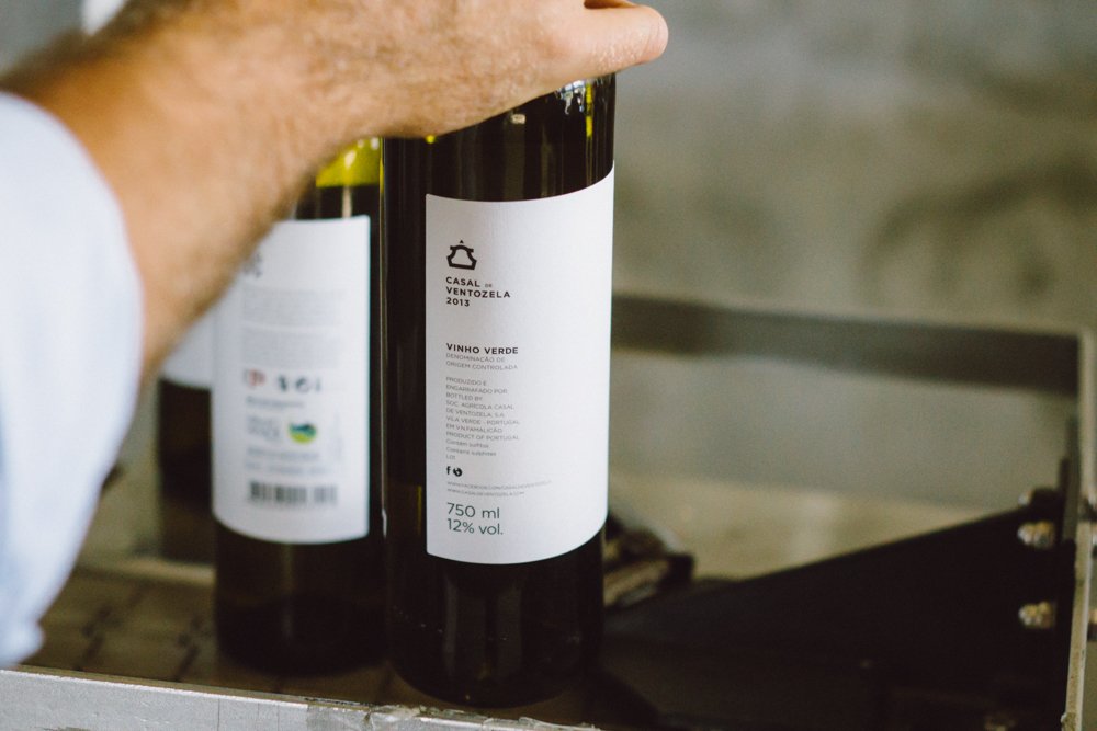 Garrafas de vinho verde com rótulos brancos sendo embaladas, uma mão segurando uma das garrafas.